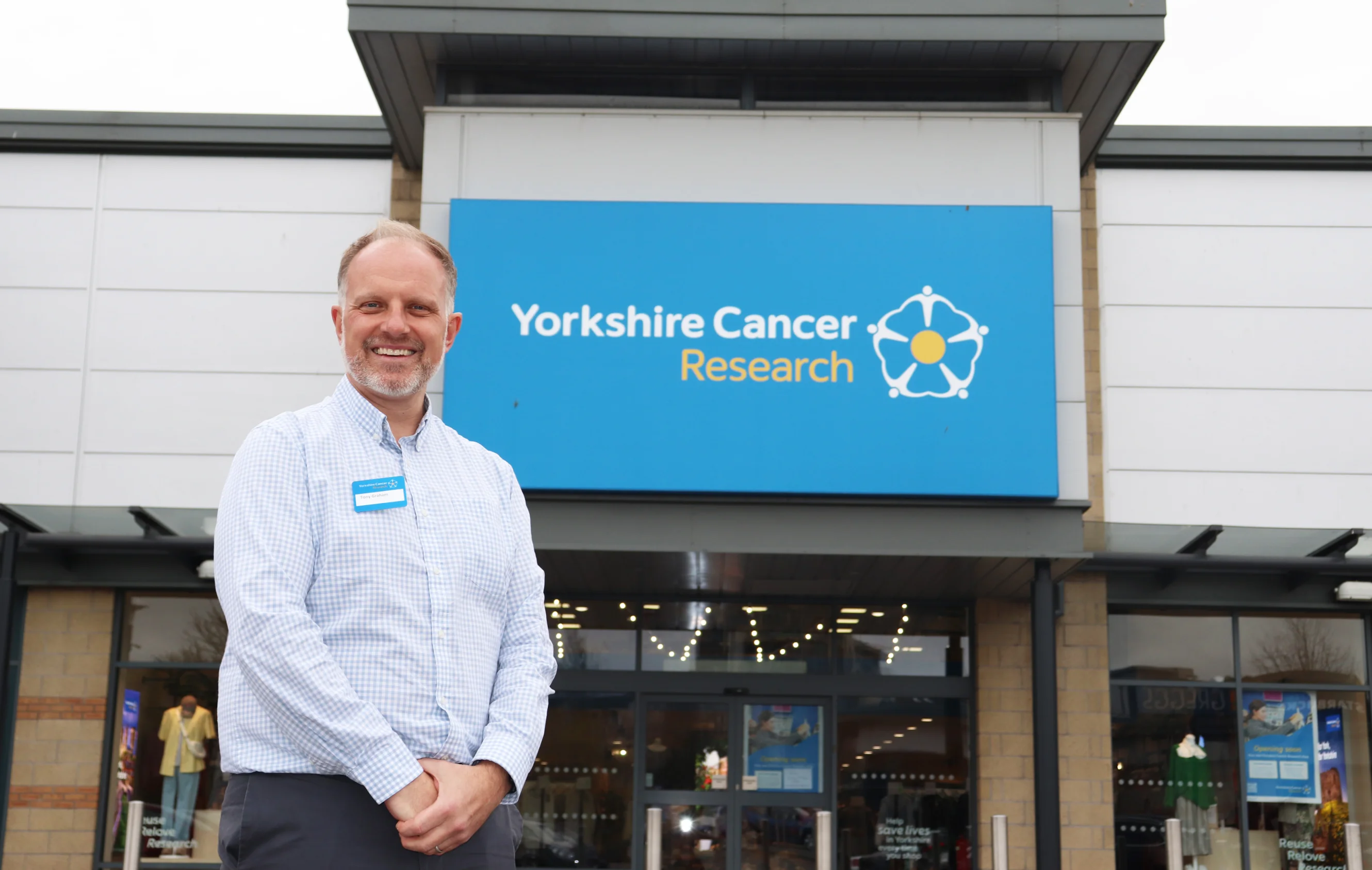 Tony is wearing a blue shirt and has a name tag on his shirt. He is standing outside the front of the shop and behind him you can see the charity's logo.