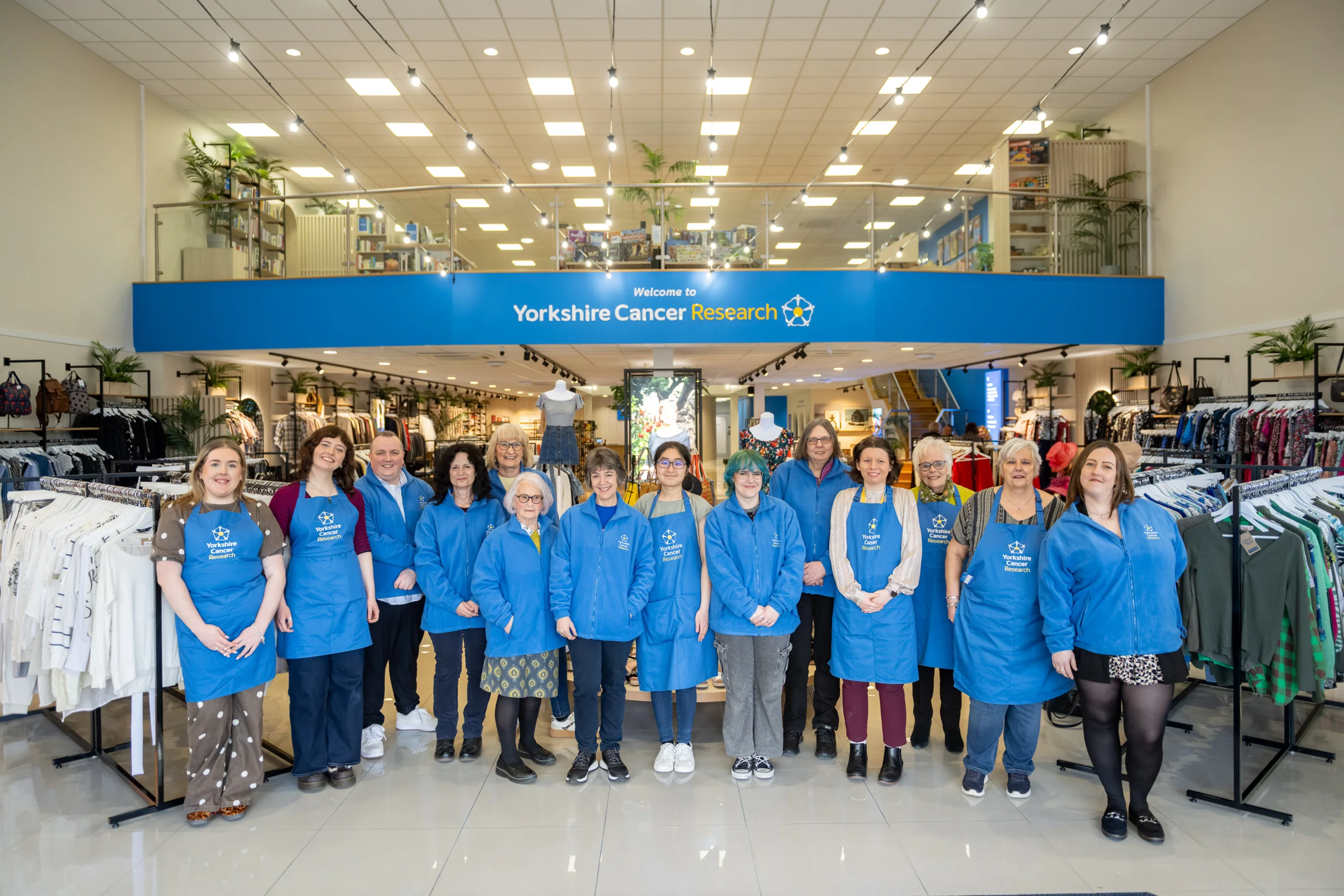 A team of staff members and volunteers all wearing blue branded "Yorkshire Cancer Research" jumpers and aprons are stood in the entrance of the shop. Behind them are clothing rails and a mezzanine showing the upper floor.