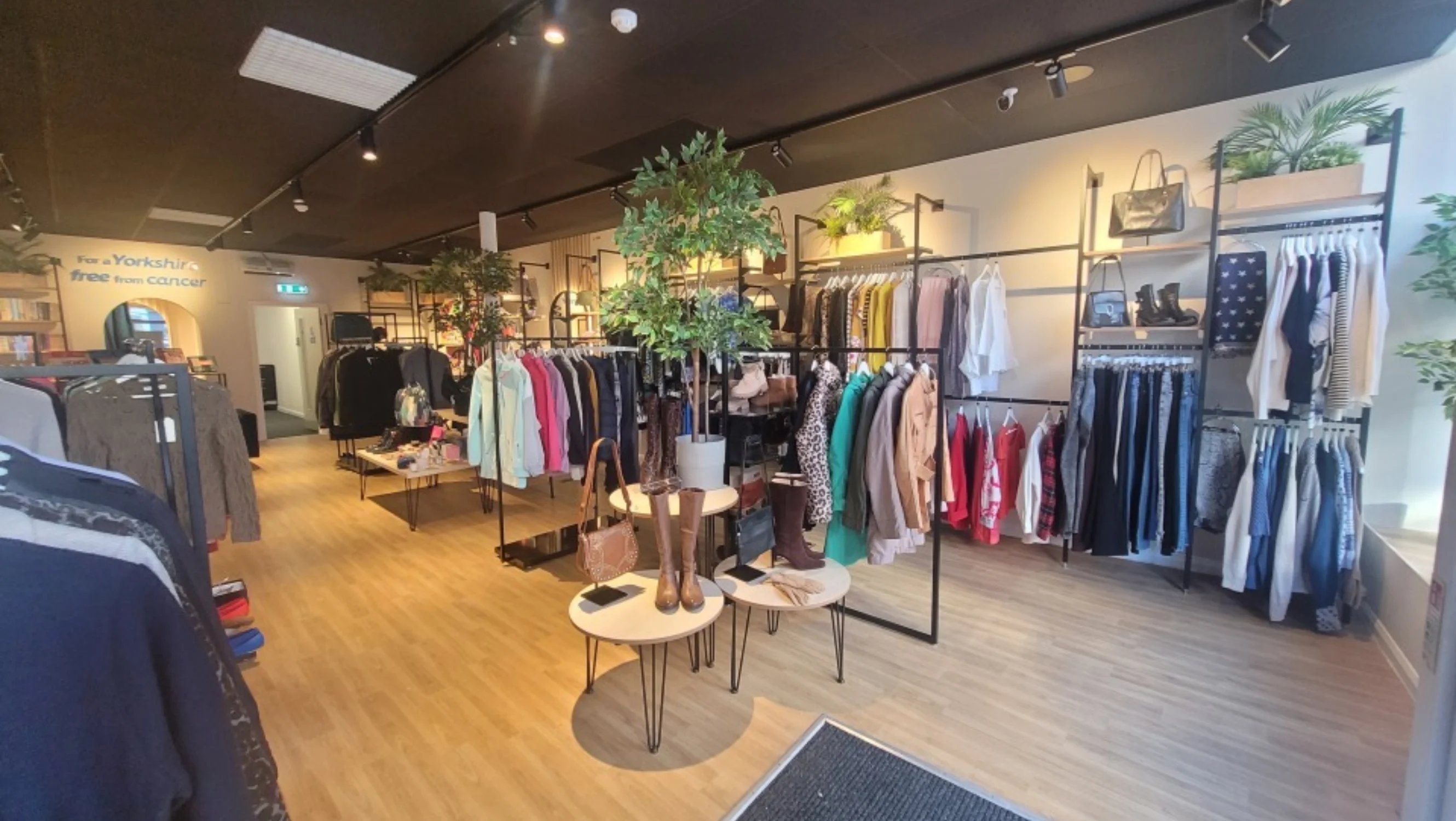 The interior of the shop. There's clothing rails, a fake plant in the centre, and spotlights on a black ceiling.