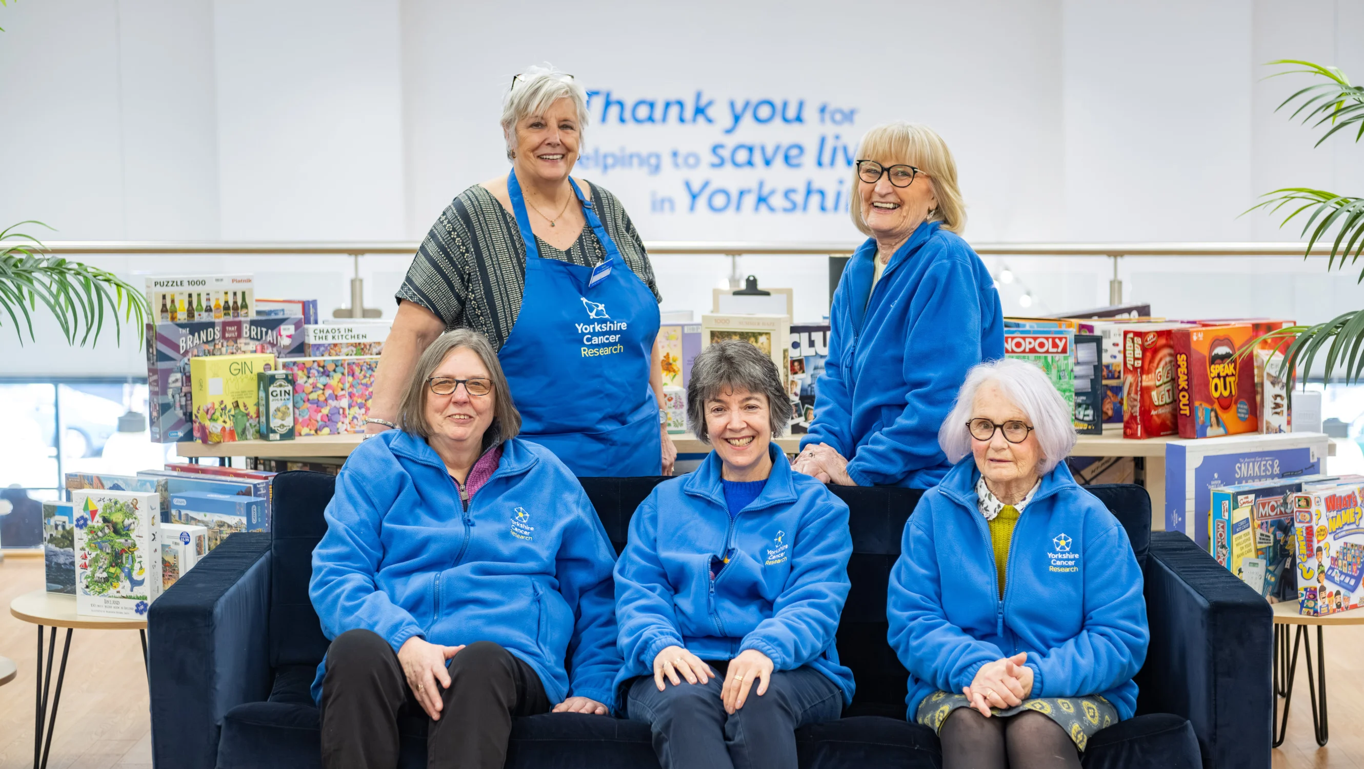 Three volunteers are sitting on a sofa in the shop. Behind them are two more volunteers leaning on the sofa. They are all smiling and wearing blue branded jumpers and aprons.