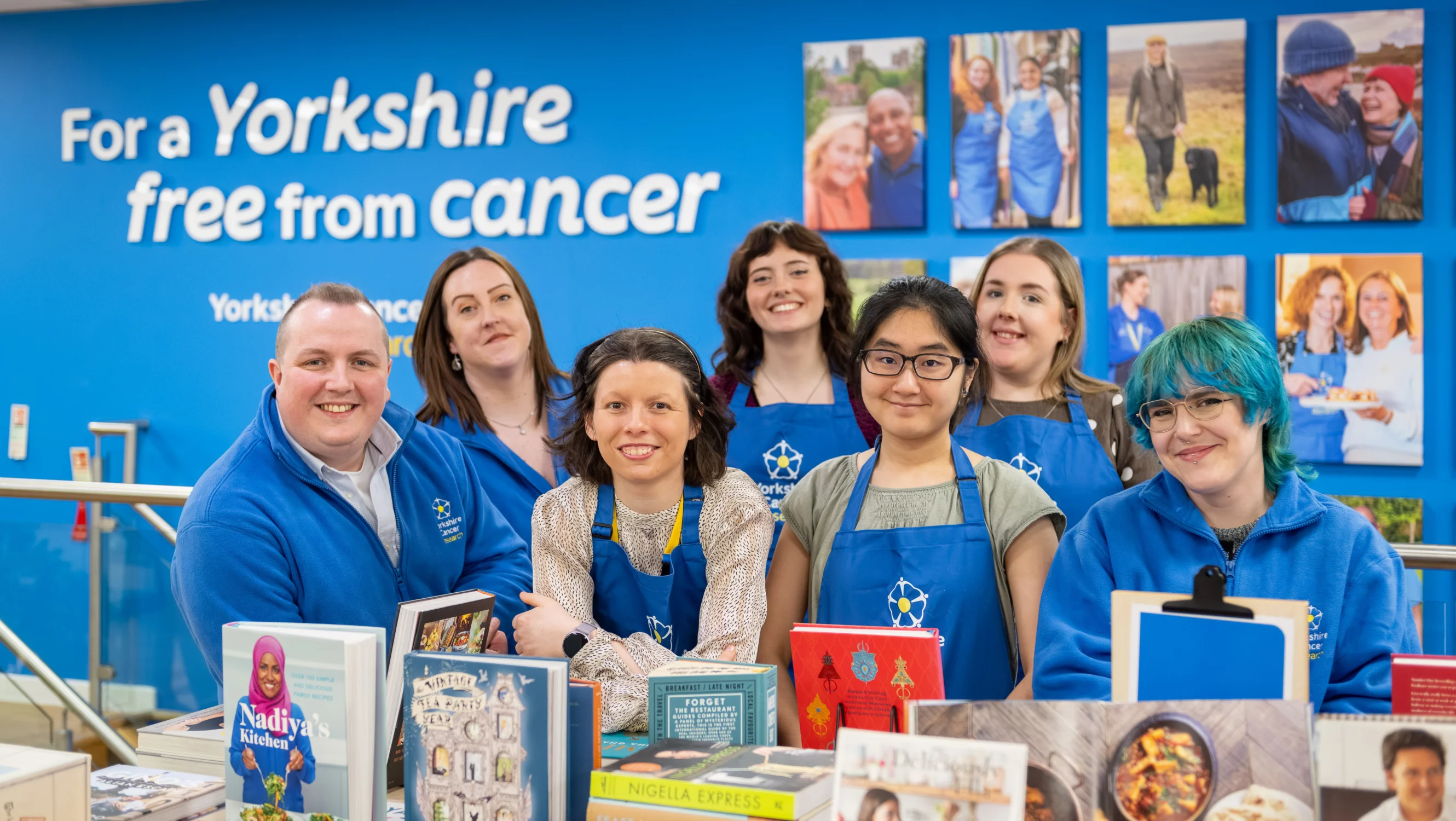 A team of seven retail staff wearing blue Yorkshire Cancer Research aprons and hoodies are stood around a table of books in the shop. Behind them is a blue wall with photos of various  volunteers and fundraisers and a sign that reads "For a Yorkshire free from cancer"