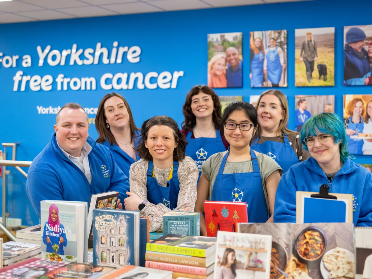A team of seven retail staff wearing blue Yorkshire Cancer Research aprons and hoodies are stood around a table of books in the shop. Behind them is a blue wall with photos of various  volunteers and fundraisers and a sign that reads "For a Yorkshire free from cancer"