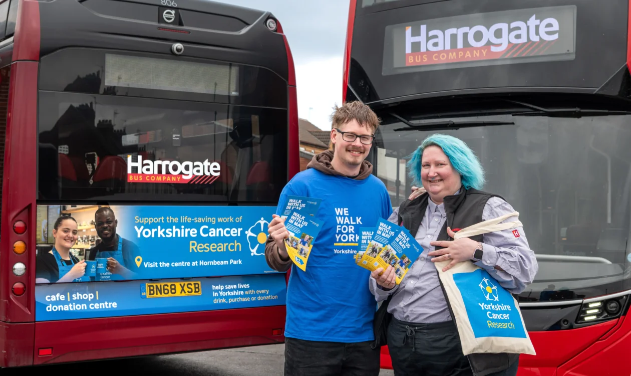 Will and Sarah stand in front of the Yorkshire Cancer Research branded bus. Will is wearing a blue We Walk for Yorkshire t-shirt and Sarah is carrying a tote bag from the charity.