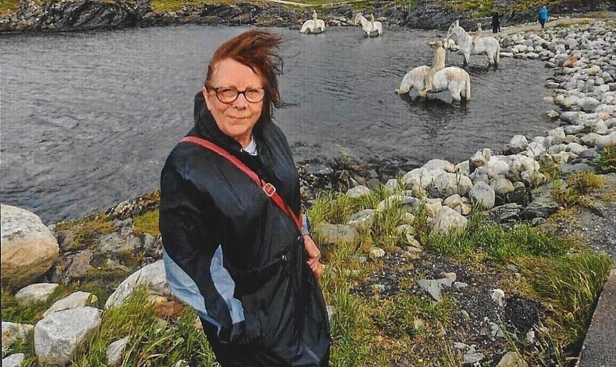 Susan is stood outside next to a lake. In the background are hills. She is wearing a coat and the weather looks windy.
