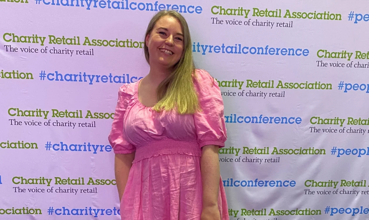 Aimee is stood in front of Charity Retail Association branding. She is smiling at the camera wearing pink