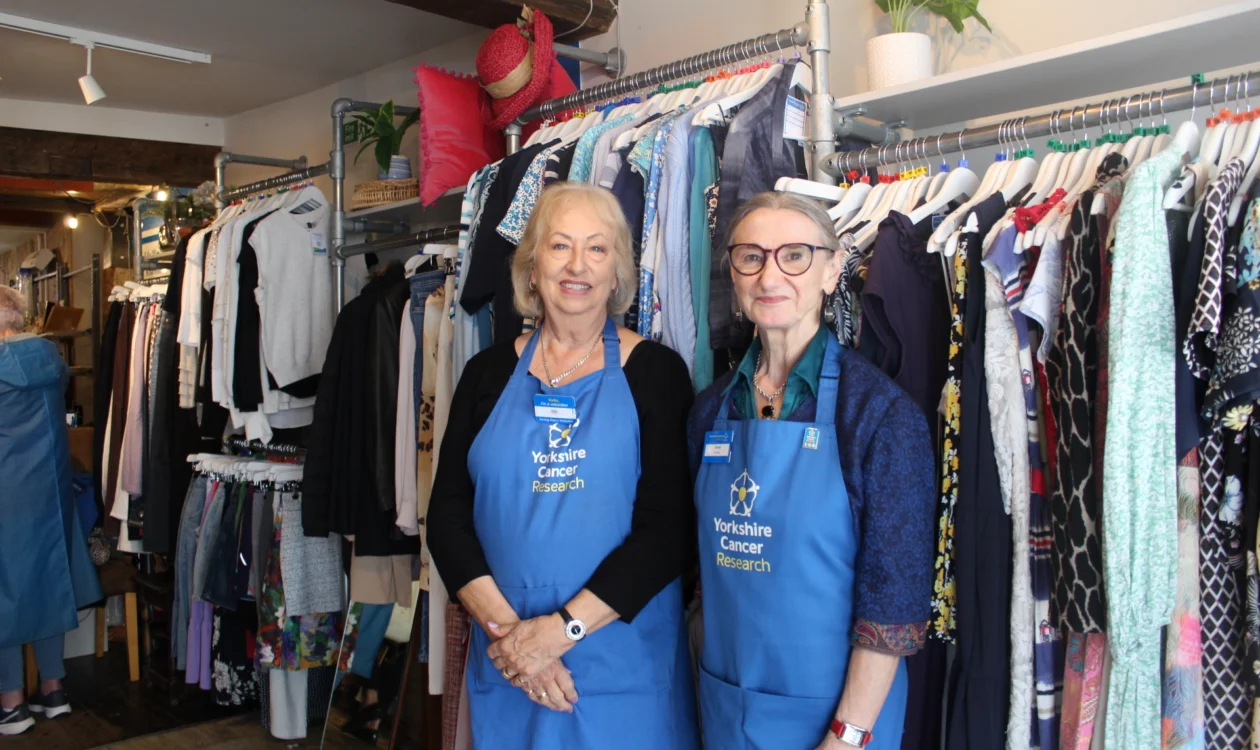 Gill and Janet are wearing blue 'Yorkshire Cancer Research' branded aprons. They are stood in the shop in front of a ladies clothing rail