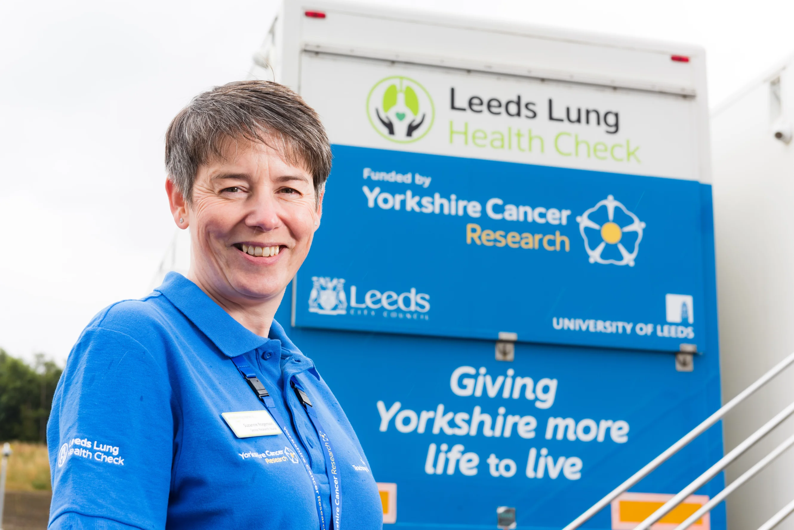 Suzanne Rogerson smiling at the camera wearing a Leeds Lung Health Check t-shirt. Behind her is a large blue screening van with the Leeds Lung Health Check logo on it.