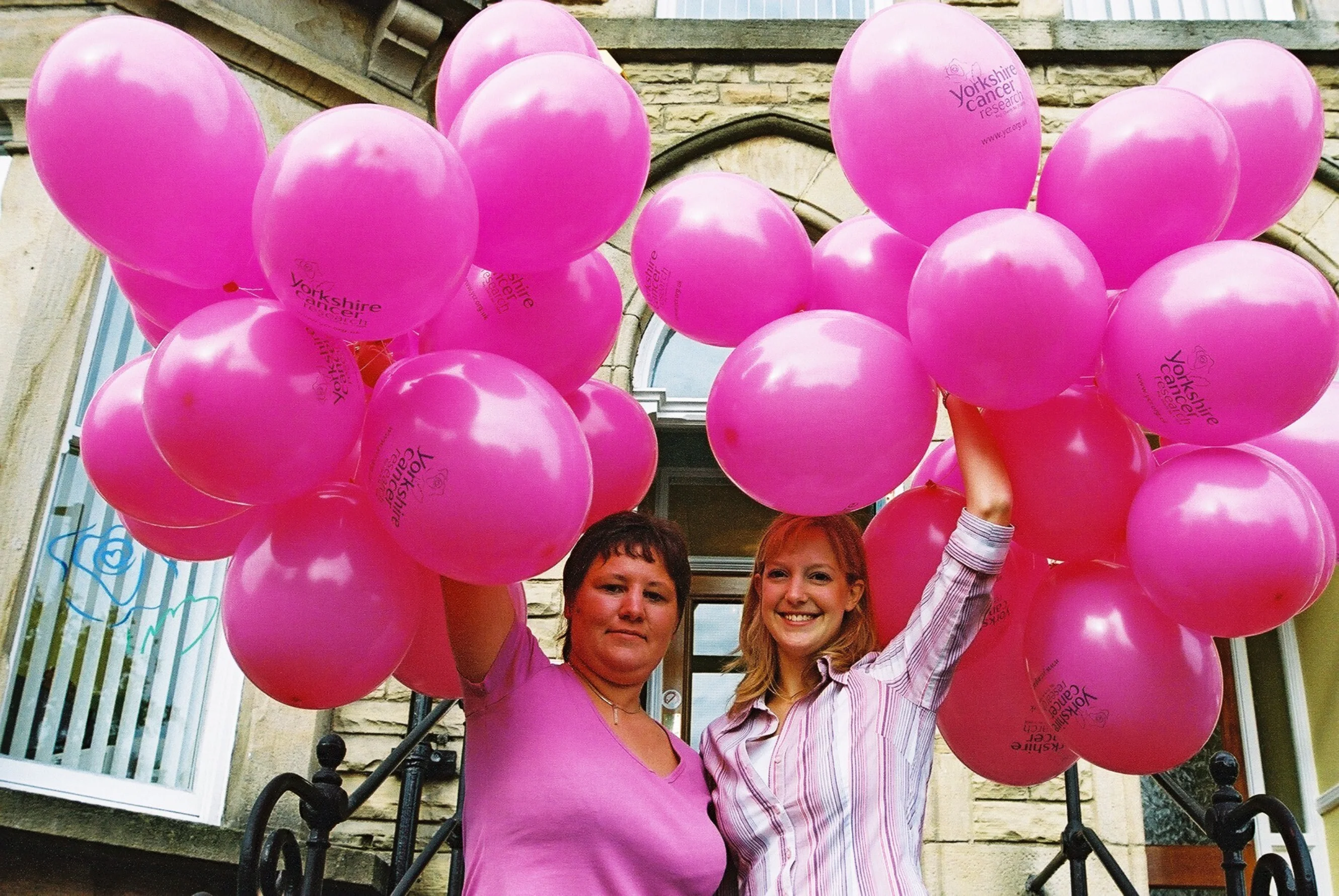 June and Rachel wearing pink clothes and each holding onto a big bunch of pink balloons, standing outside a townhouse.