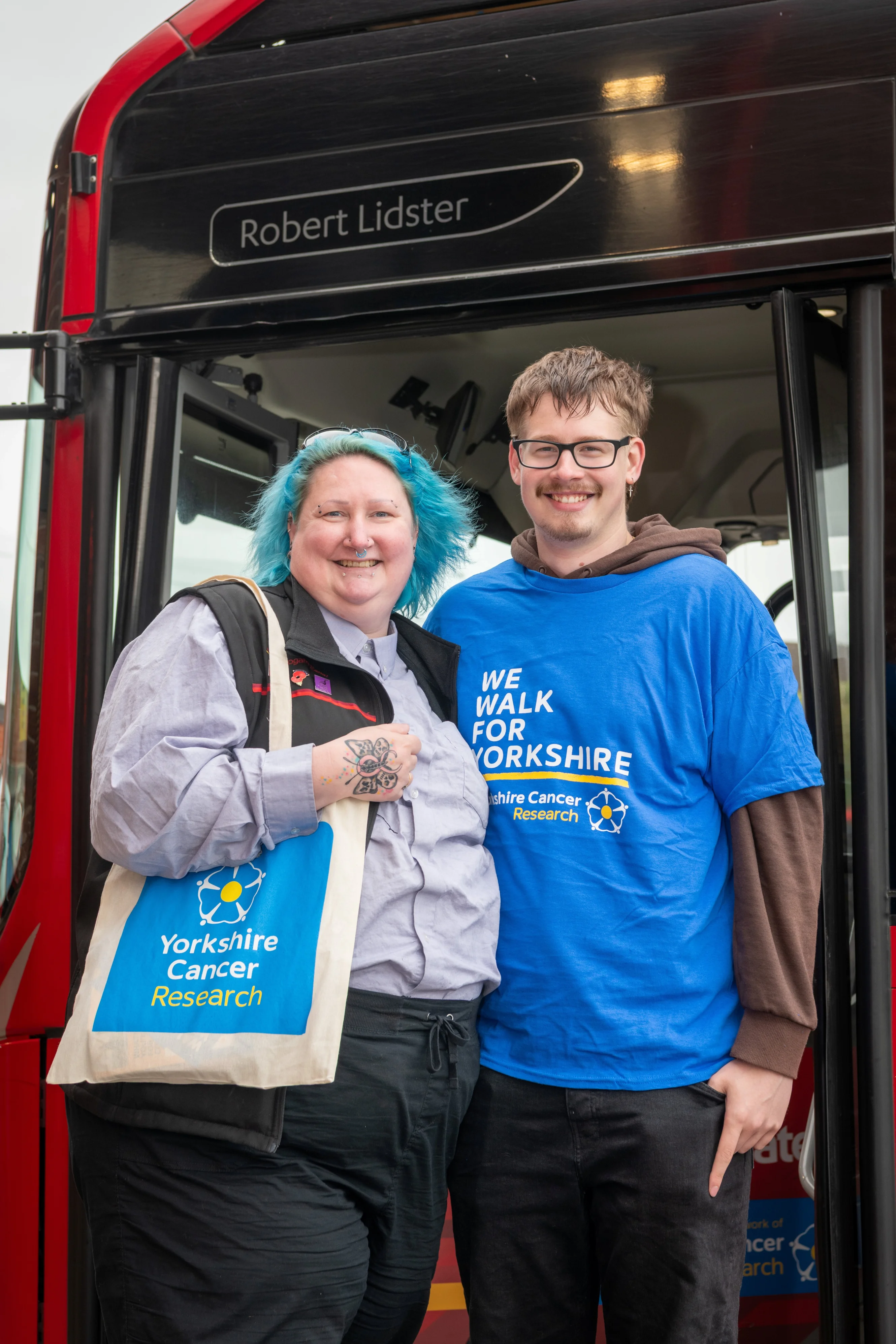 Sarah is standing on the left wearing a grey bus driver uniform and carrying a Yorkshire Cancer Research tote bag. Will is standing on the right wearing a Yorkshire Cancer Research t-shirt. They are in front of a bus.
