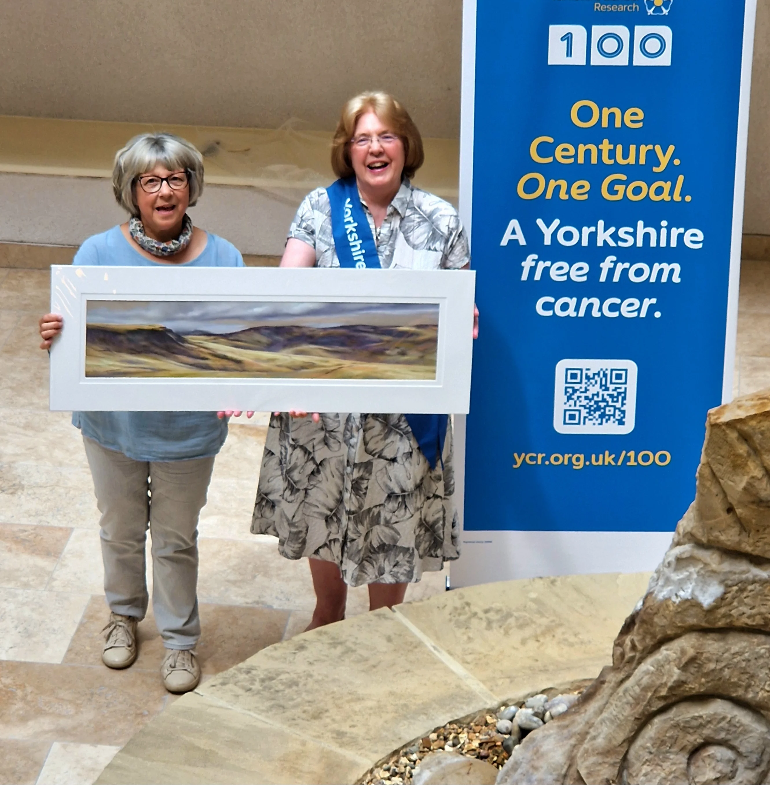 Lucia Smith alongside Katy Bell Yorkshire Cancer Research Ilkley Committee member with CLASS ACT III. They are both smiling while holding the painting.