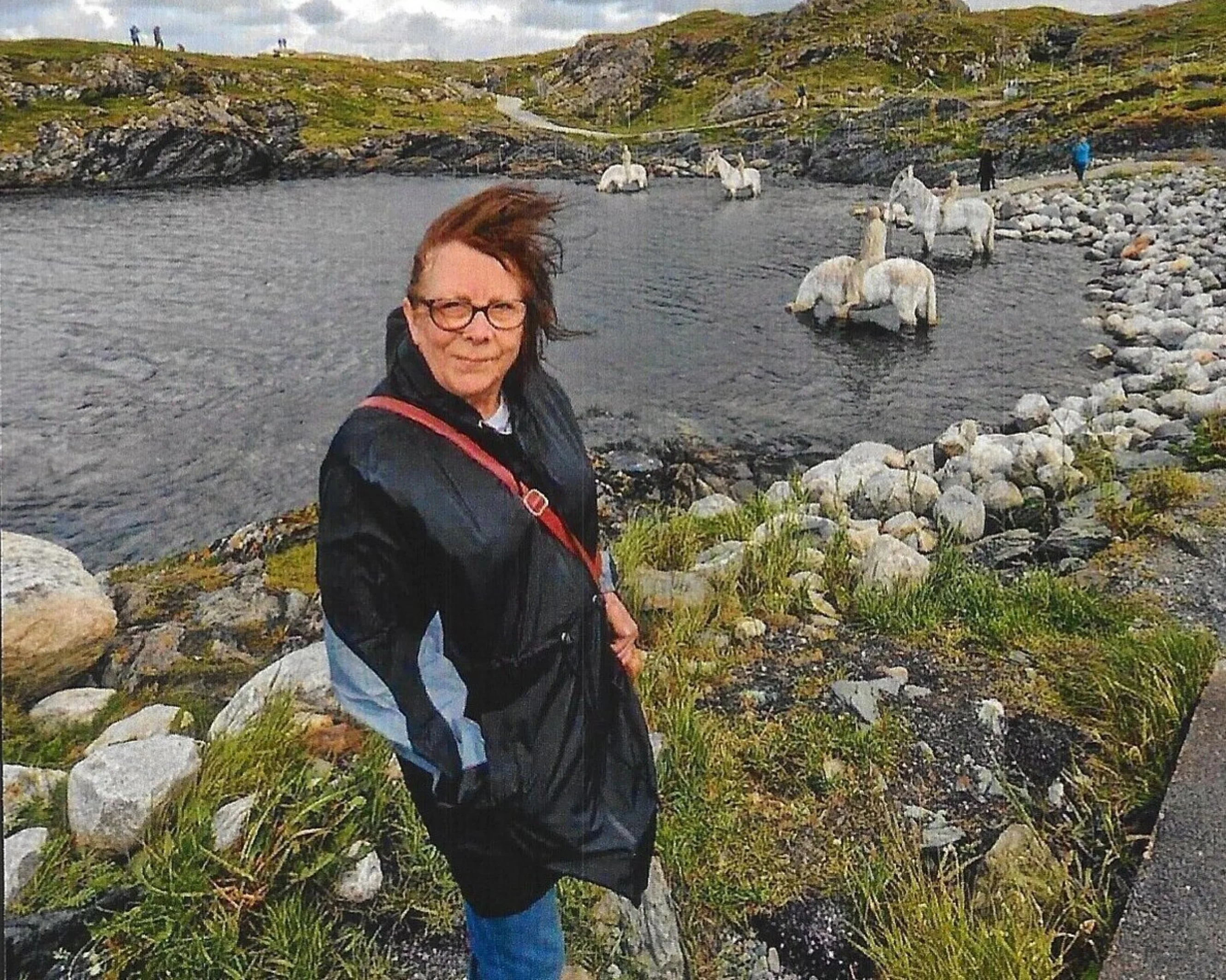 Susan is stood outside next to a lake. In the background are hills. She is wearing a coat and the weather looks windy.