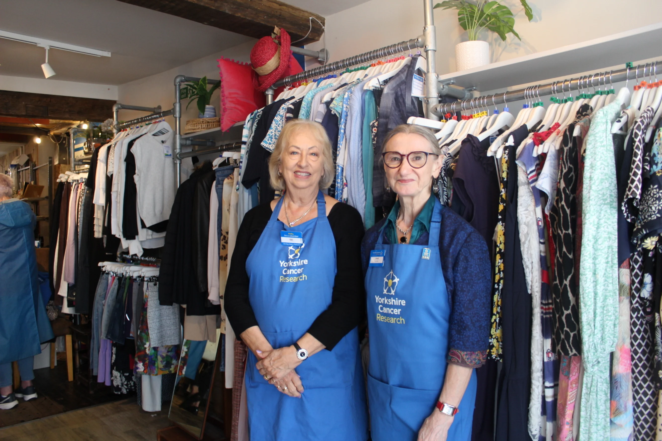 Gill and Janet are wearing blue 'Yorkshire Cancer Research' branded aprons. They are stood in the shop in front of a ladies clothing rail