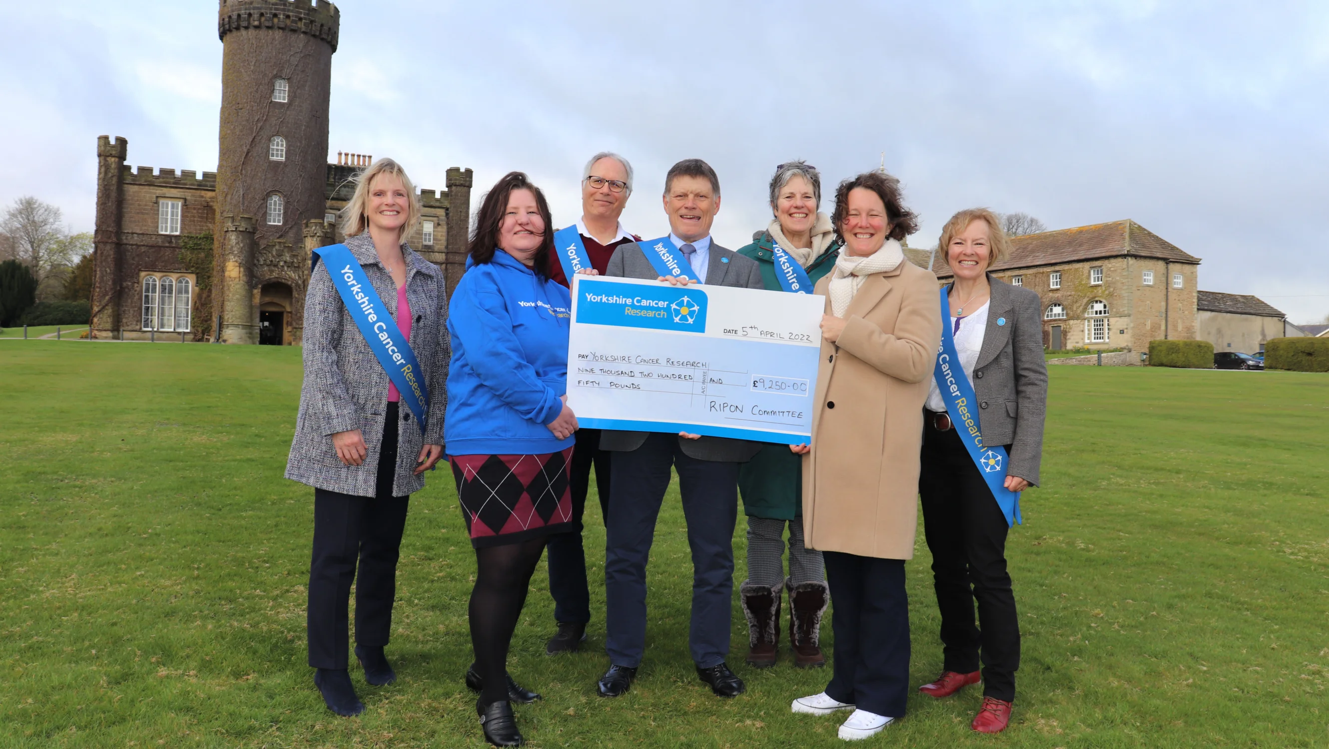 A group of fundraisers handing over a big cardboard cheque to June. The fundraisers are wearing blue Yorkshire Cancer Research sashes and they are outdoors on a grass field.