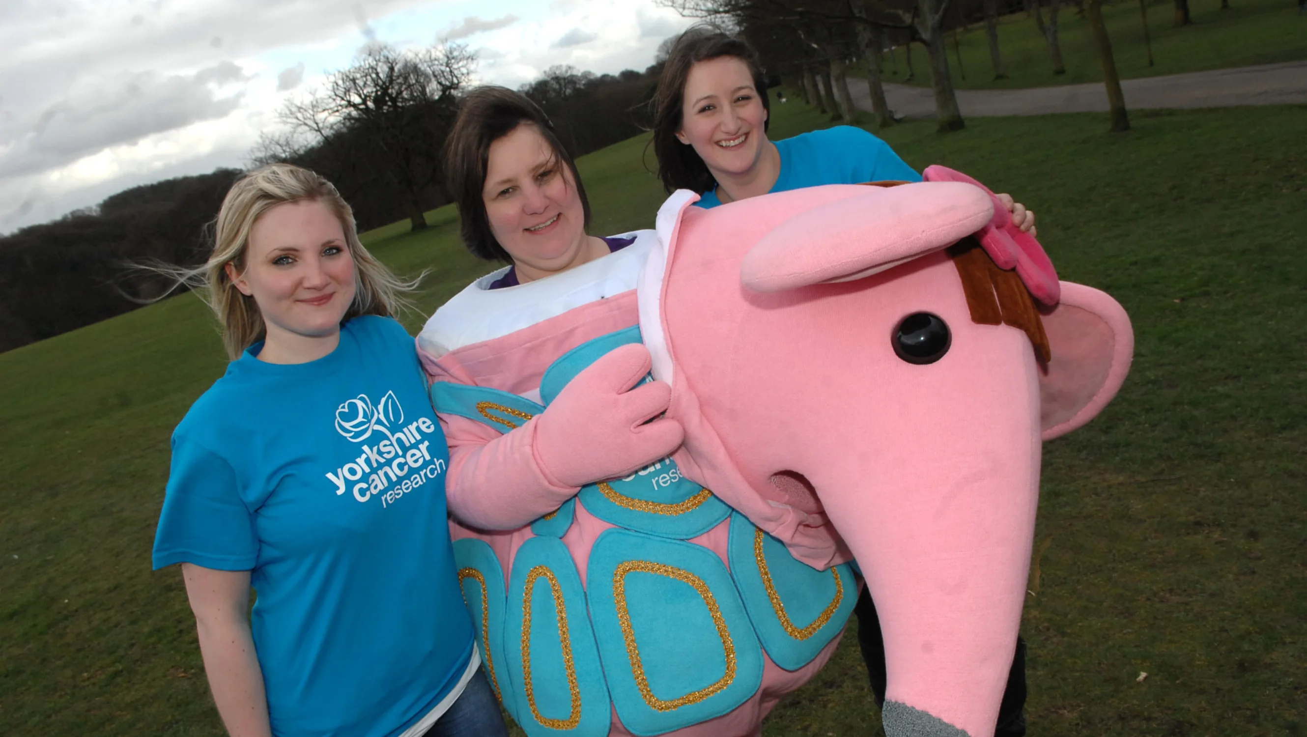 Three ladies on a windy field, the two on the sides are wearing blue Yorkshire Cancer Research t-shirts and June is in the middle in a Clangers costume.