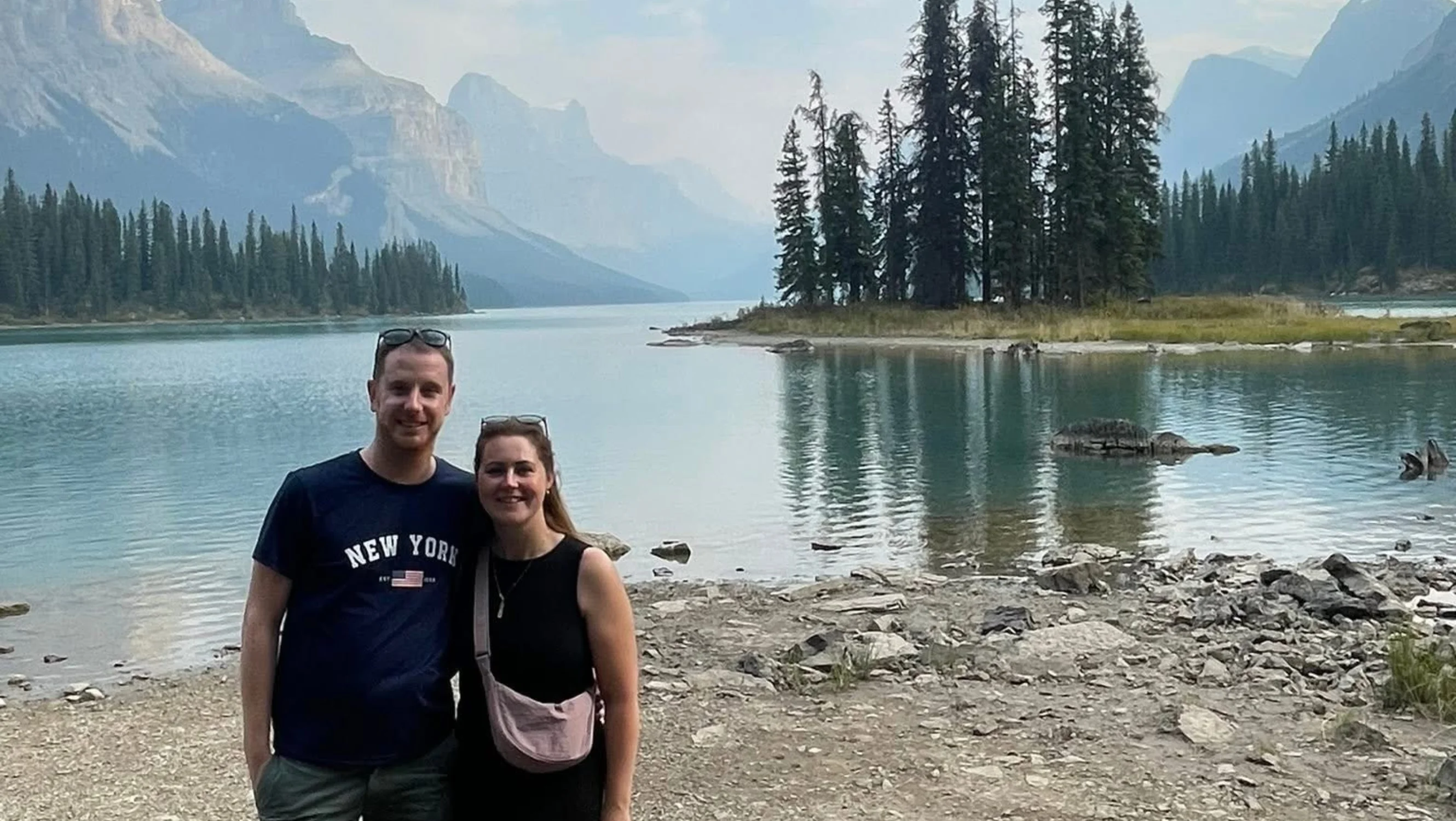 A man and woman pose in front of a body of water and mountains in the background