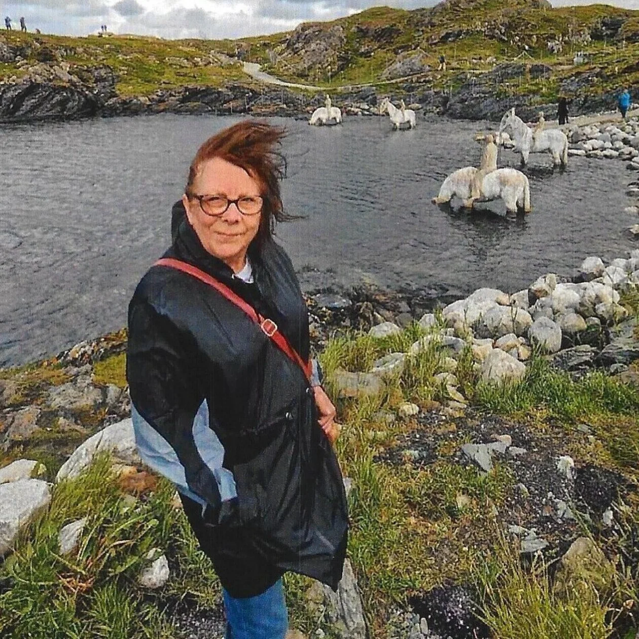 Susan is stood outside next to a lake. In the background are hills. She is wearing a coat and the weather looks windy.