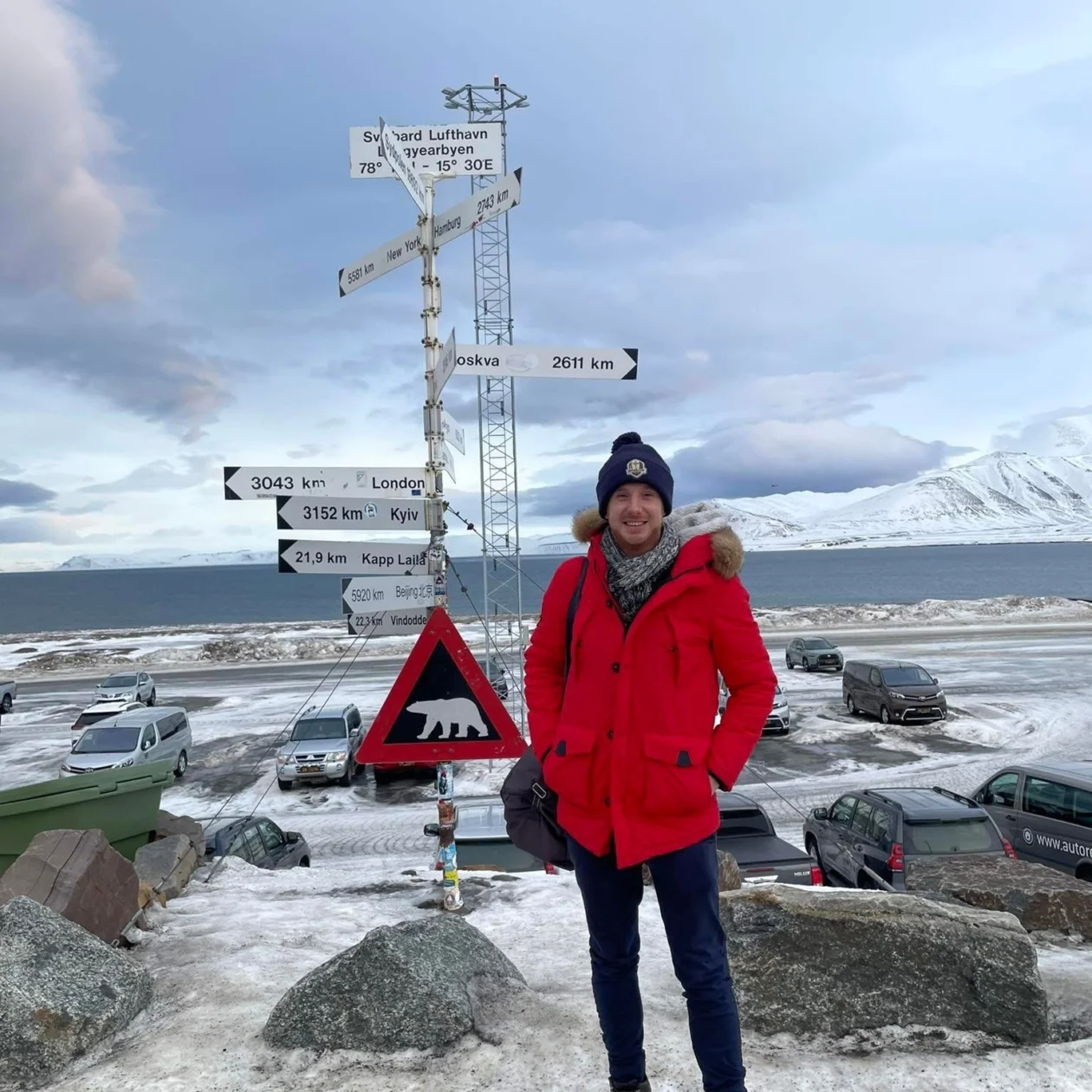 A man wrapped up in winter clothes stands in front of wayfinder signs for different countries