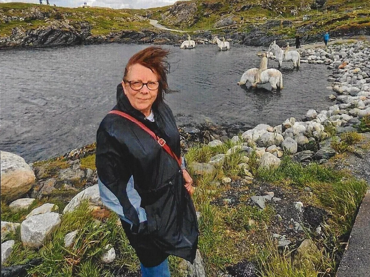 Susan is stood outside next to a lake. In the background are hills. She is wearing a coat and the weather looks windy.