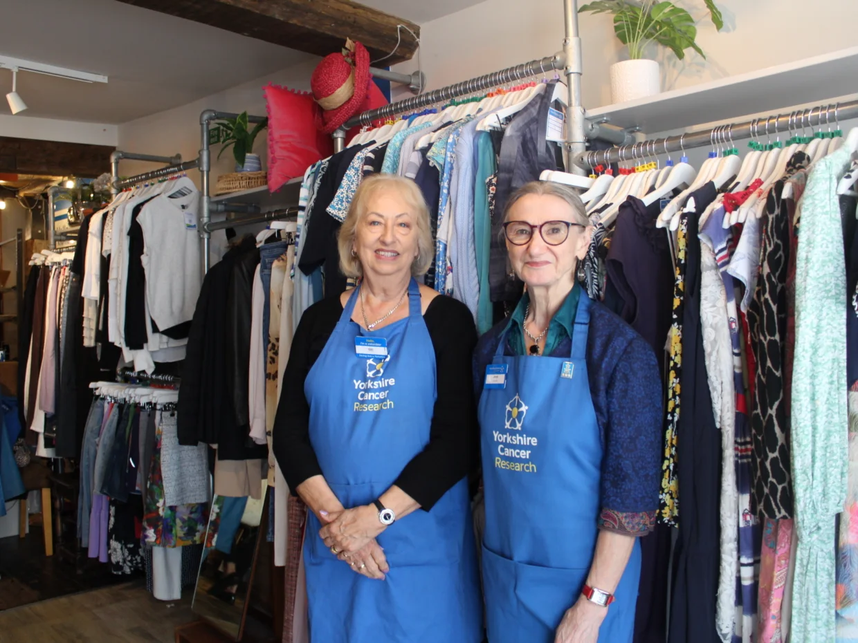 Gill and Janet are wearing blue 'Yorkshire Cancer Research' branded aprons. They are stood in the shop in front of a ladies clothing rail