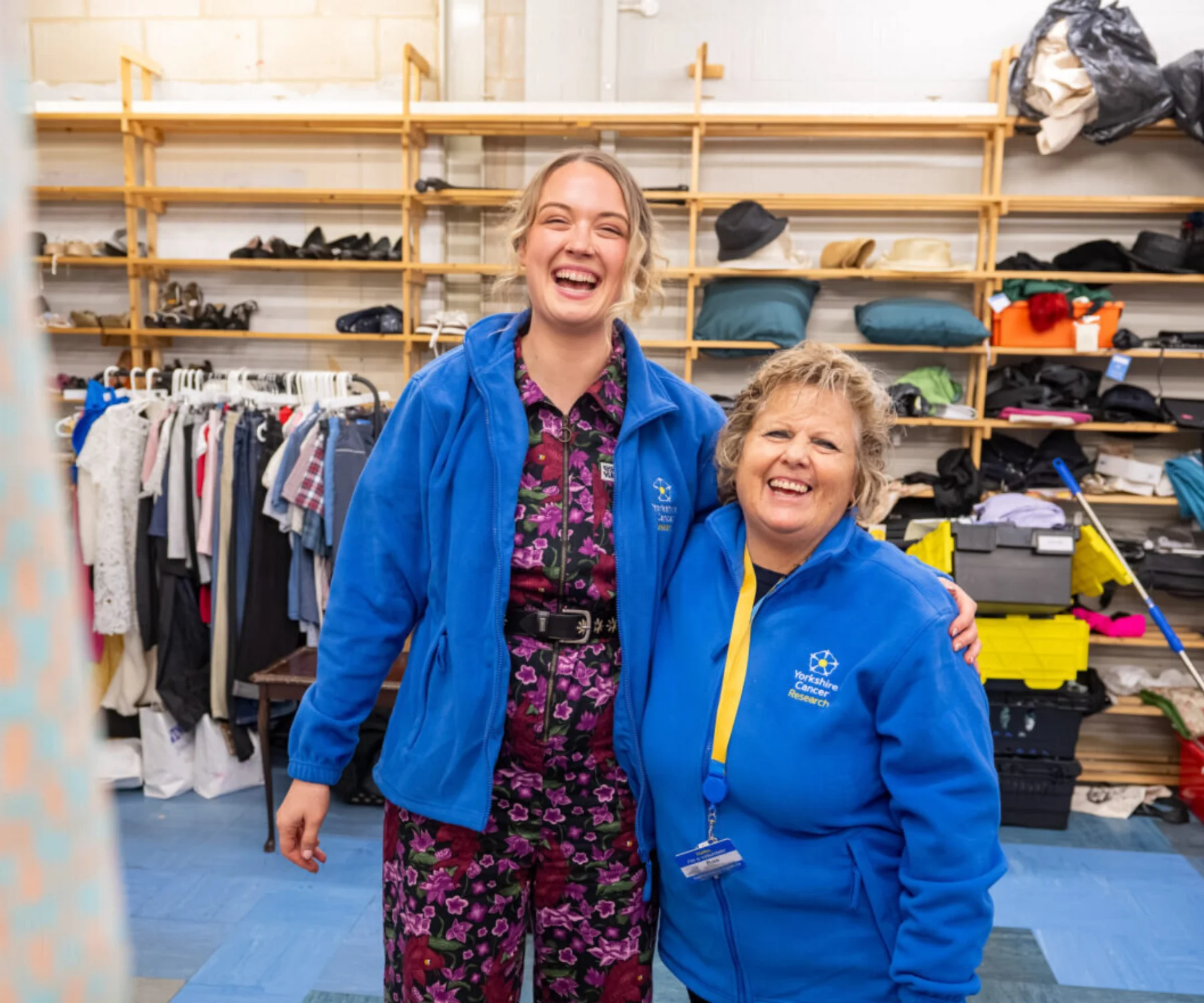 Two ladies are stood in the back of a shop. Behind there are shelves for storage and rails of clothing. They are both wearing blue "Yorkshire Cancer Research" fleeces.