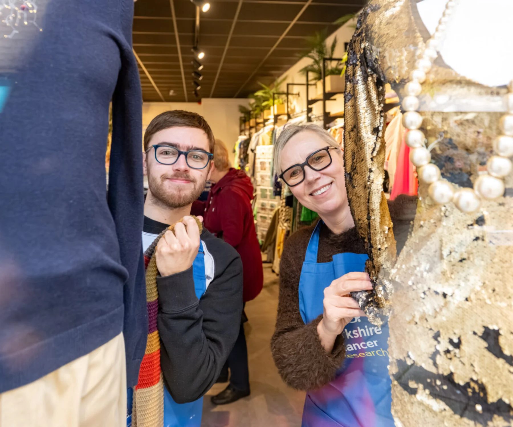 Two people are standing behind mannequins in a shop window display. They are both leaning in to get into the shot. The mannequins are in sparkly clothes and the shop shelves are visible behind them.