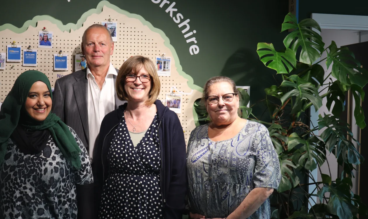 Four people are standing in front of a dark green wall with a map of Yorkshire on it. Above the map is a wall decal that reads "Here for Yorkshire" There are three ladies and one gentleman smiling at the camera.