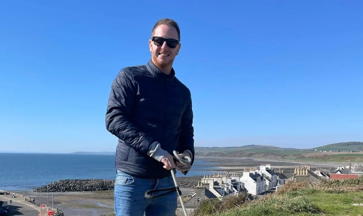 Stephen is outside standing by a cliff edge on a coastline. The sky is blue and the grass a vibrant green. He is wearing sunglasses and a navy coat.