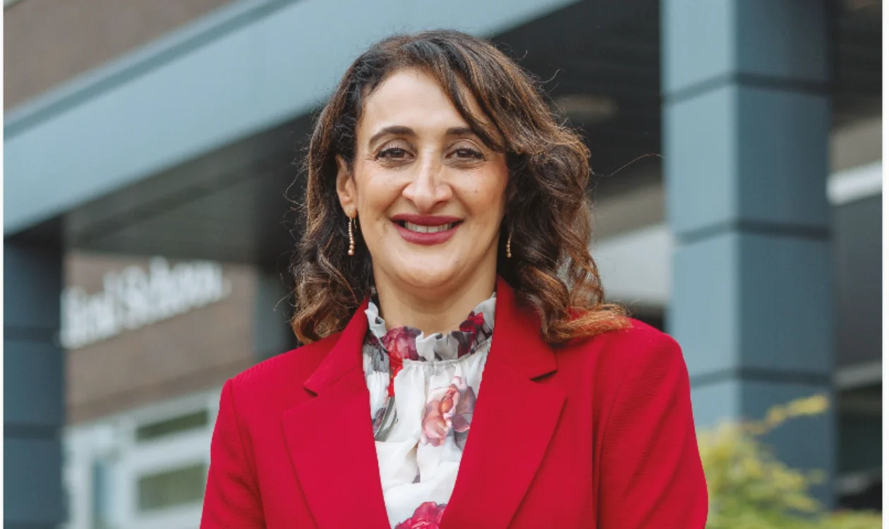 Munitta is standing in front of an academic building. She is wearing a bold red suit and patterned blouse.