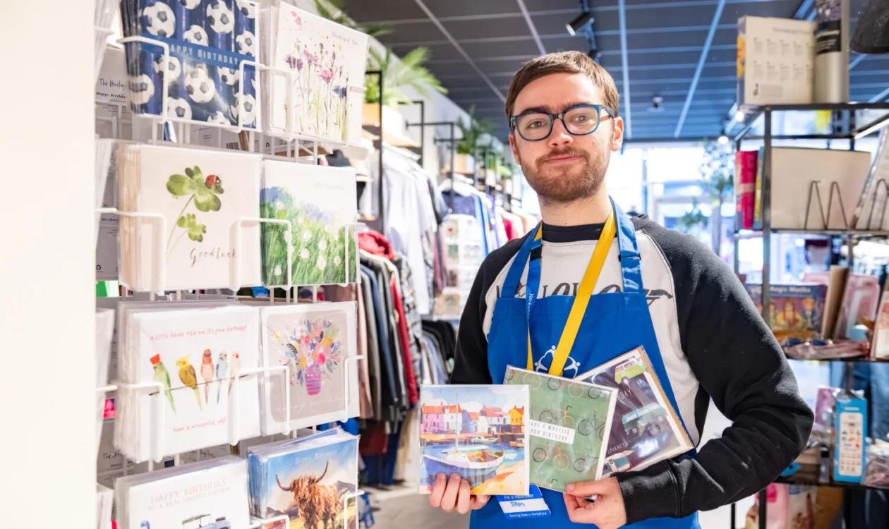 A volunteer is wearing a blue Yorkshire Cancer Research apron and is holding two packs of greetings cards. To the left of him is a tall card stand. Behind him is the rest of the Halifax shop.