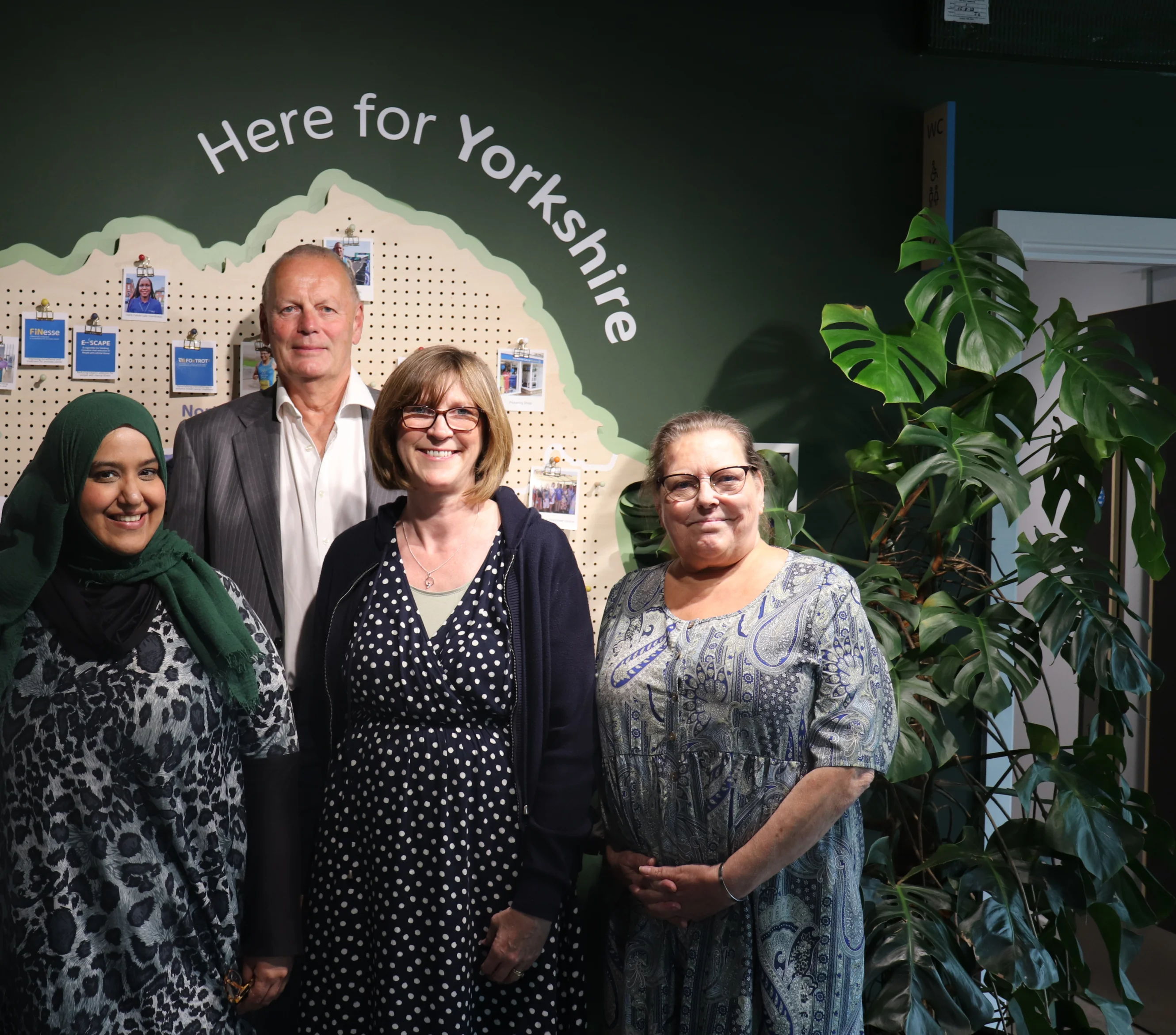 Four people are standing in front of a dark green wall with a map of Yorkshire on it. Above the map is a wall decal that reads "Here for Yorkshire" There are three ladies and one gentleman smiling at the camera.
