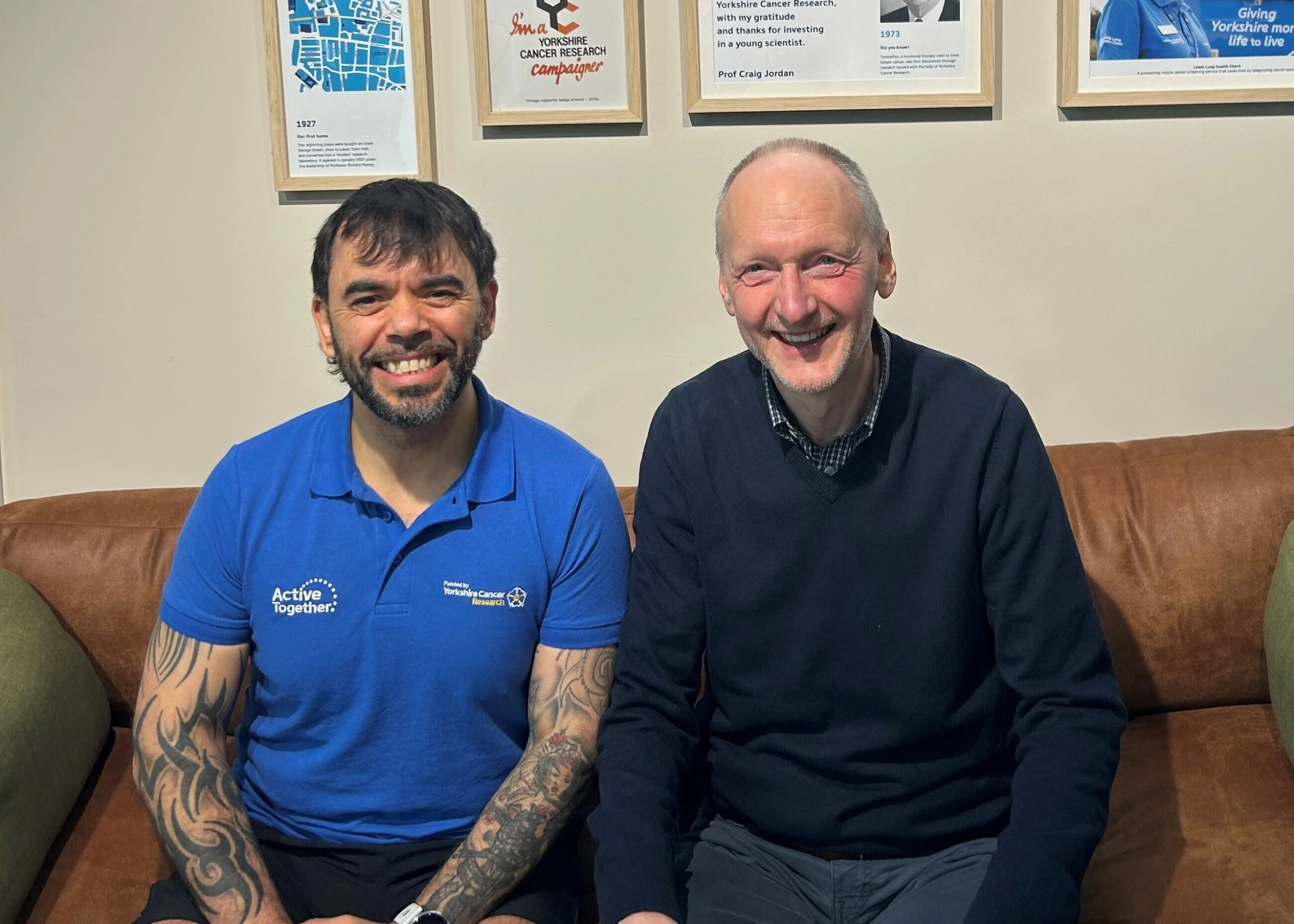 Barry is sitting on a brown leather sofa next to a fitness instructor who is wearing a blue Active Together t-shirt. Both men are smiling at the camera.