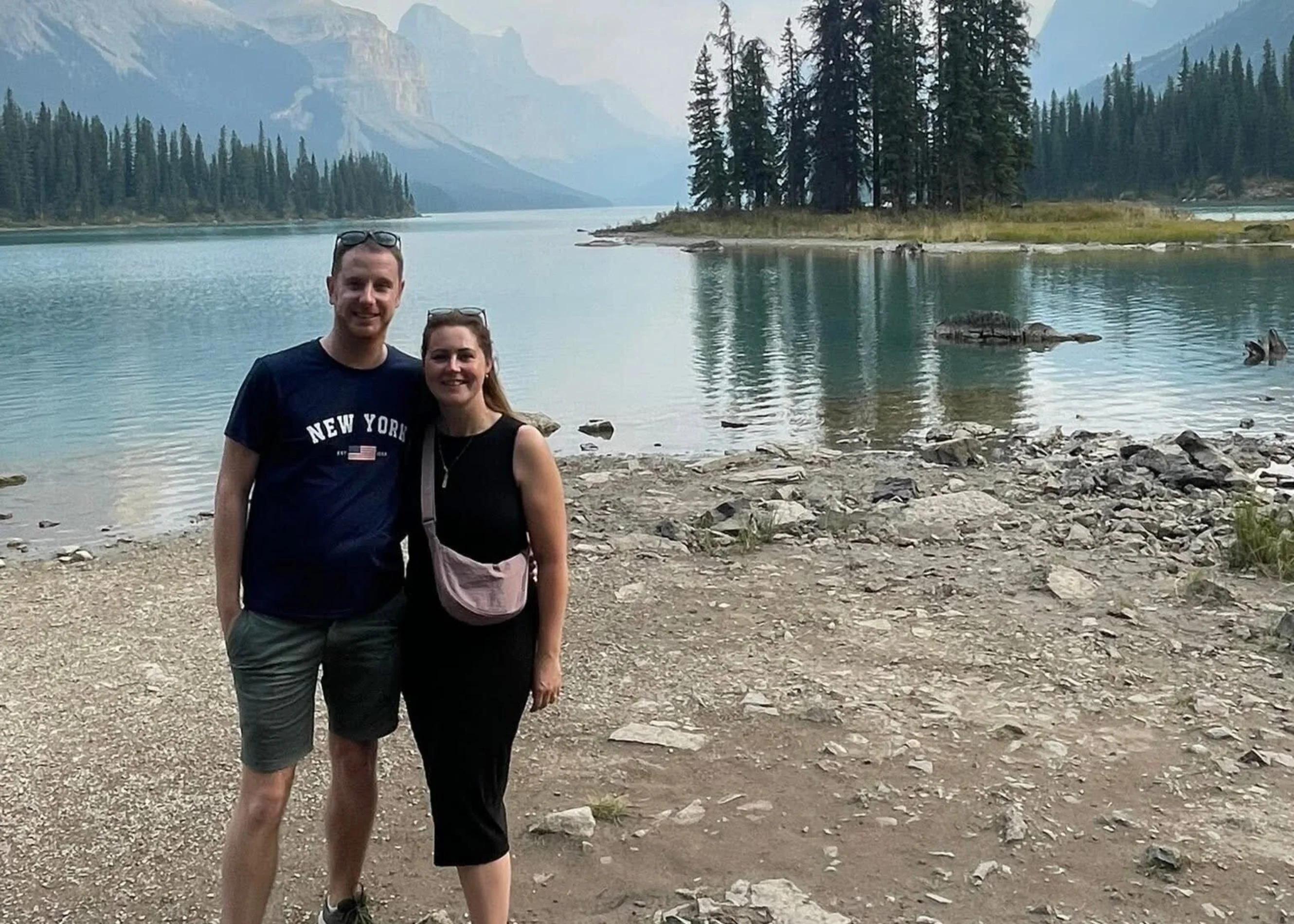 Stephen and his wife are standing on the shores of an impressive lake on their travels. Behind them are large mountains and tall trees. The water in the lake is a striking icey blue.