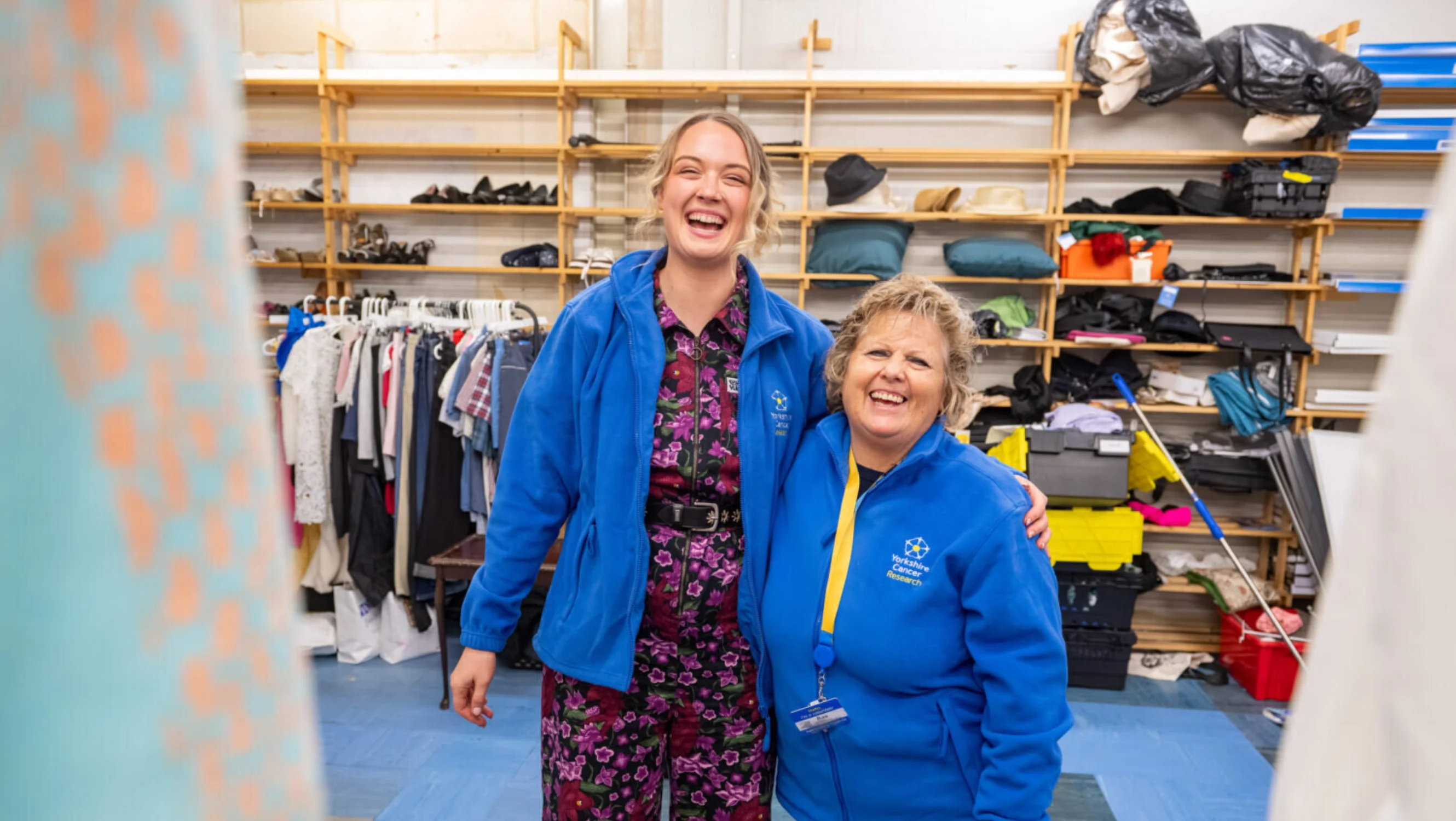 Two ladies are stood in the back of a shop. Behind there are shelves for storage and rails of clothing. They are both wearing blue "Yorkshire Cancer Research" fleeces.