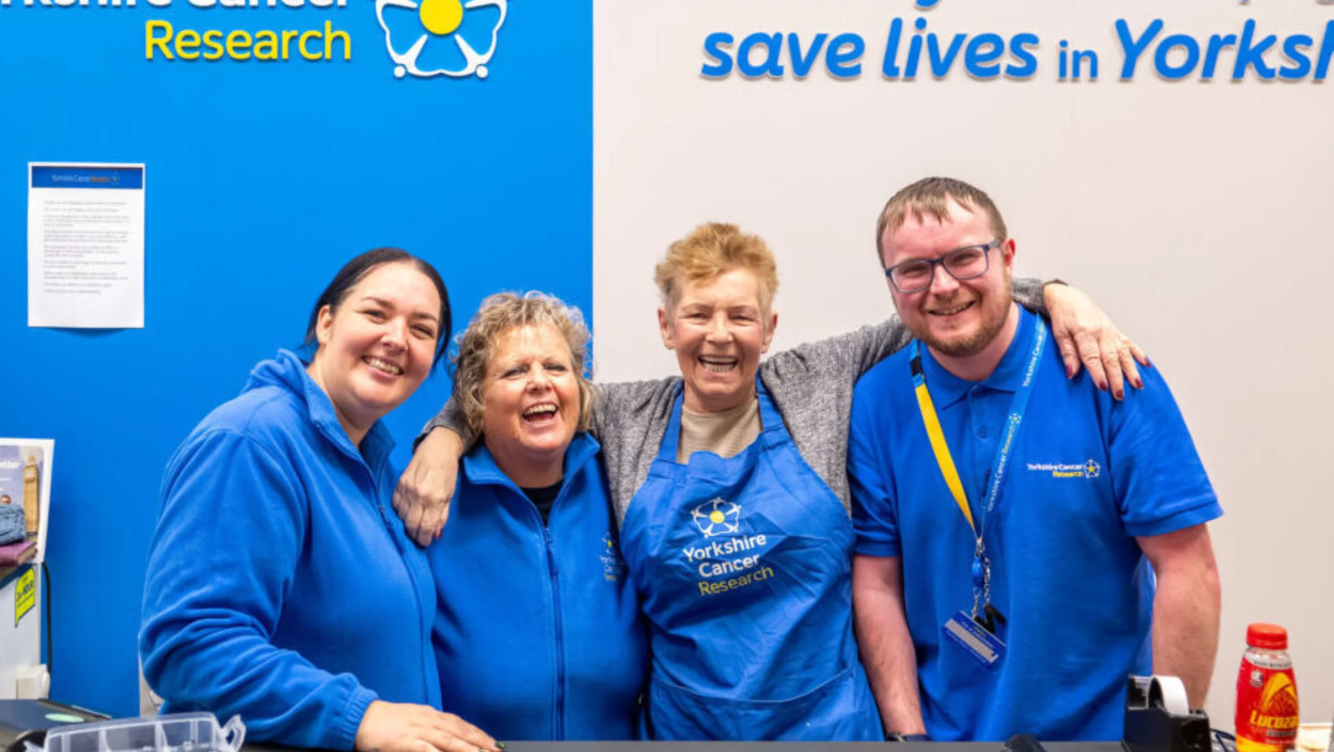 Four people are standing by a till wearing blue branded charity clothes. They all appear very happy. Behind them is a blue wall with the charity's logo and a sign that reads "Thank you for helping to save lives in Yorkshire"