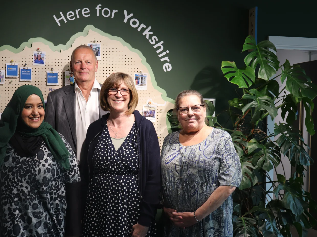 Four people are standing in front of a dark green wall with a map of Yorkshire on it. Above the map is a wall decal that reads "Here for Yorkshire" There are three ladies and one gentleman smiling at the camera.