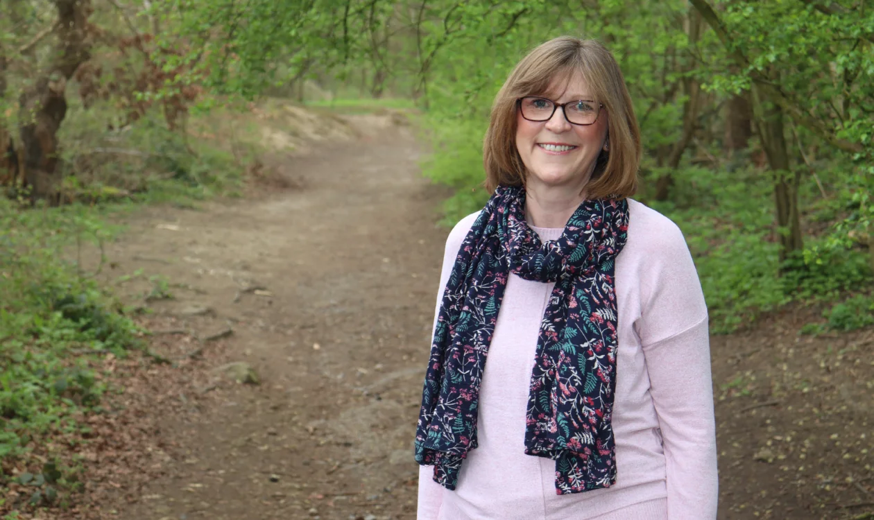 Tracey is standing in front of some trees and smiling at the camera, She's wearing a pink top and a floral scarf.