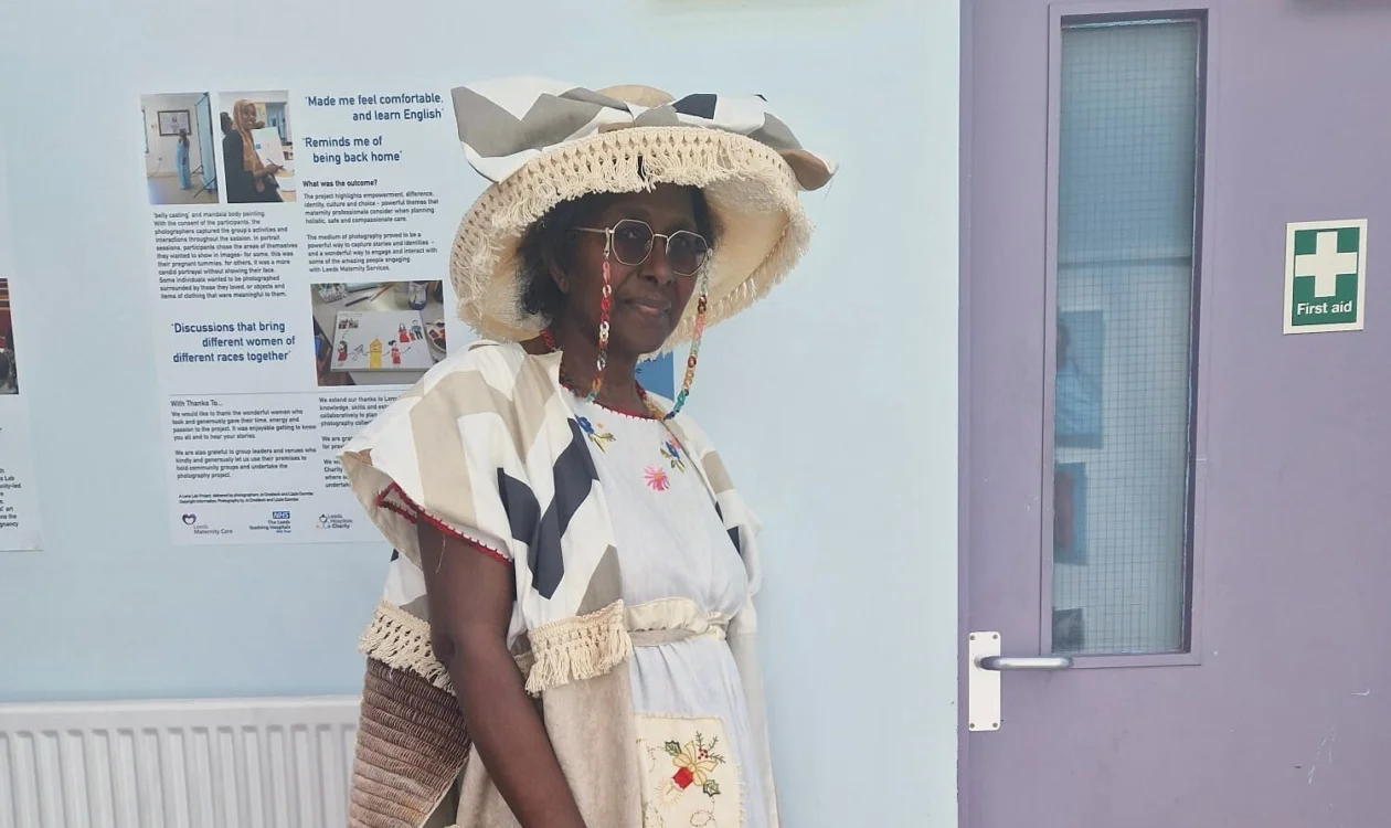 Gloria in the outfit Goodness designed - a beige hat, a long white dress with embroided details, and a long beige jacket.