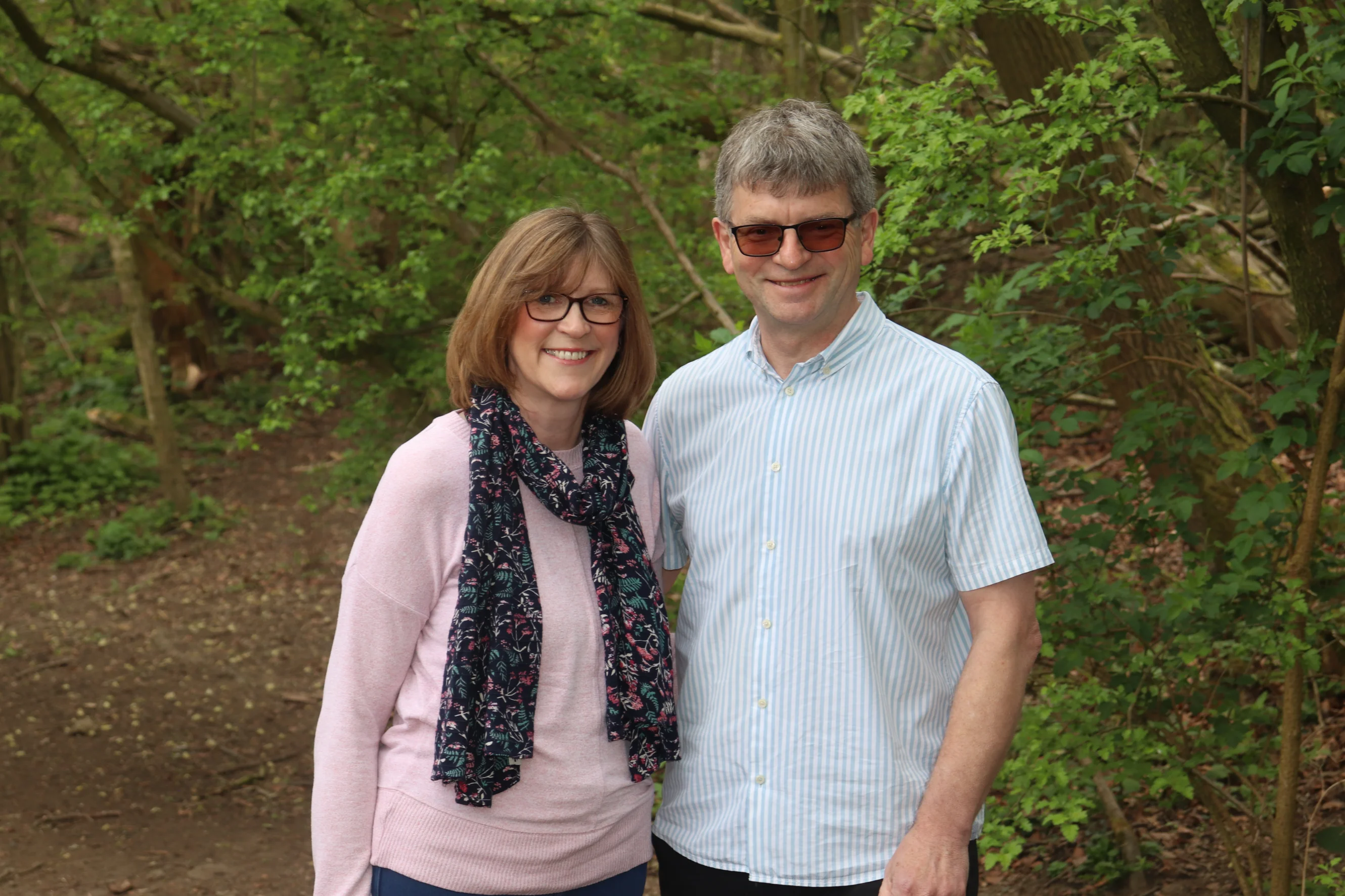 Tracey and her husband stand in front of some trees and smile at the camera.