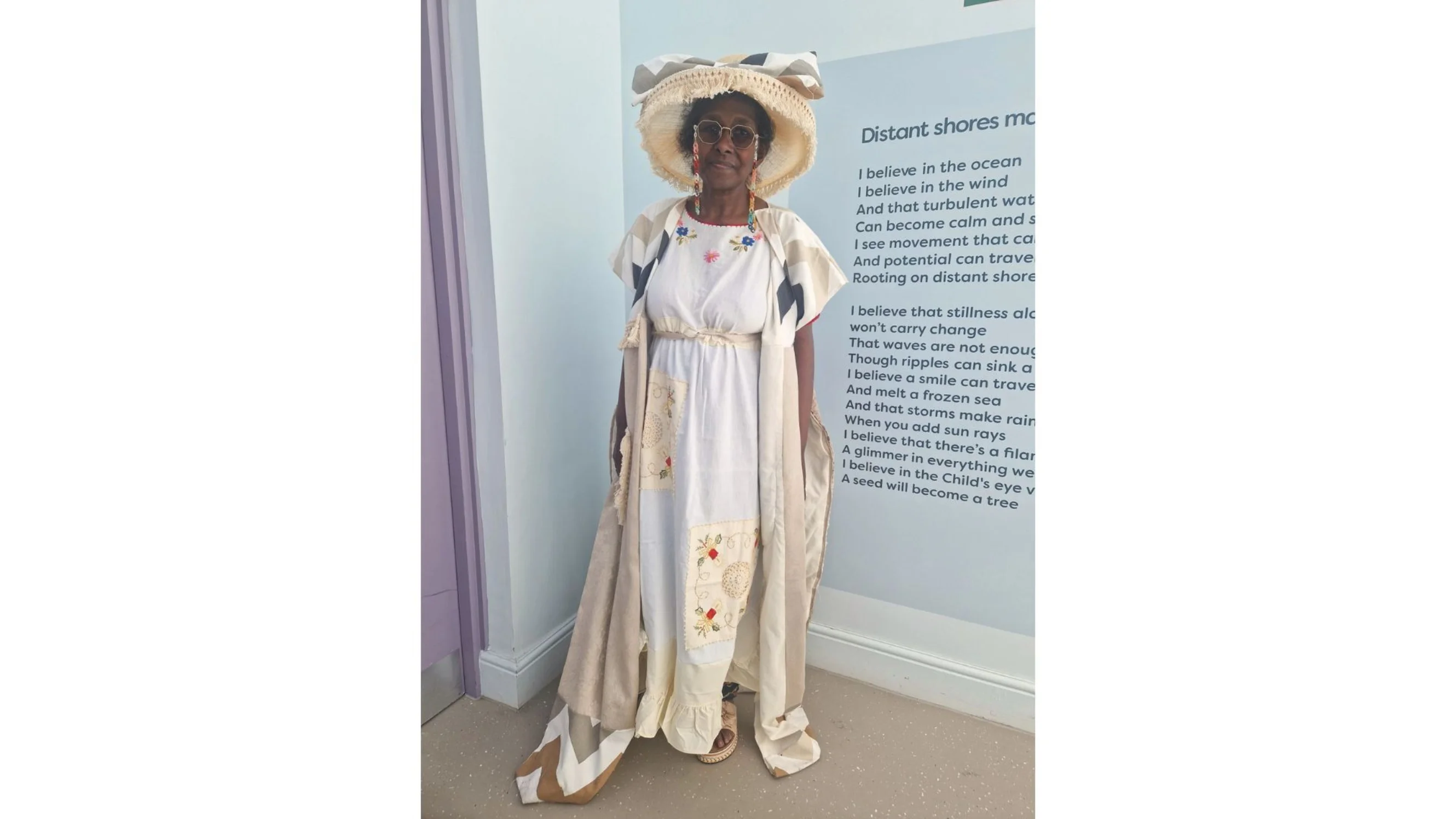 Gloria in the outfit Goodness designed - a beige hat, a long white dress with embroided details, and a long beige jacket.