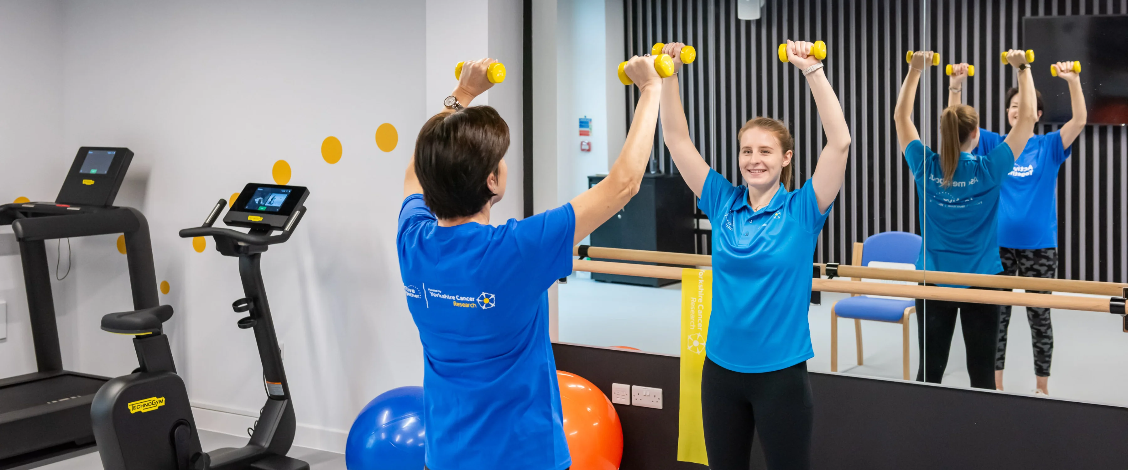 A woman exercising with a weights in a gym with a fitness instructor