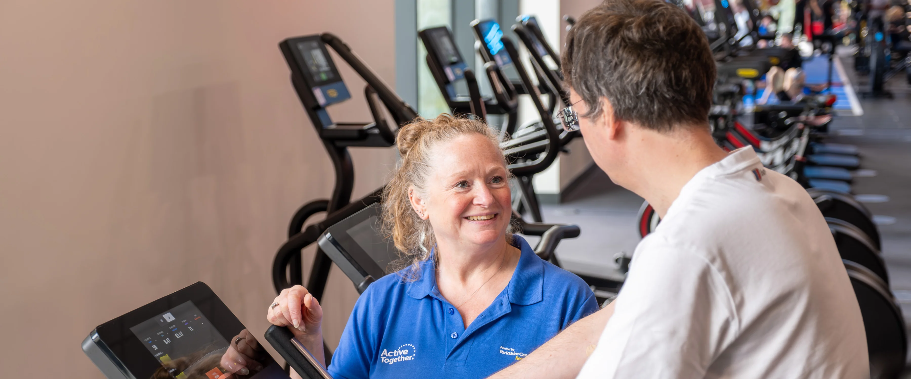 A person exercising on a bike supported by a member of staff in blue