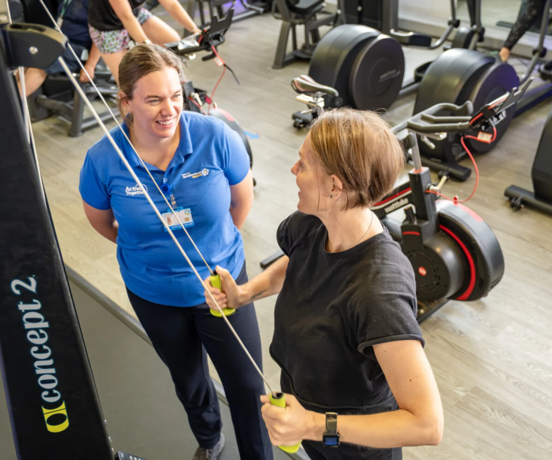 A woman wearing black exercising in a gym supported by a woman wearing blue