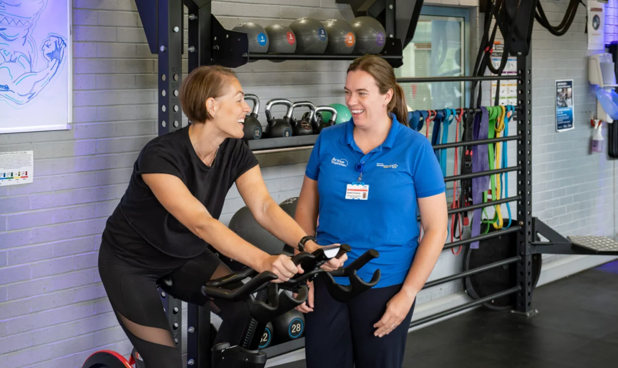 A person exercising on a bike in a gym with a member of staff dressed in blue