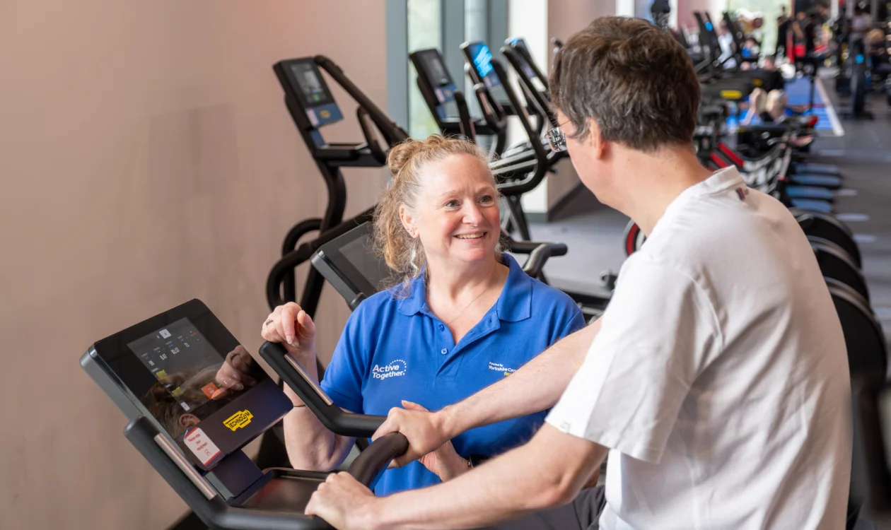 A person exercising on a bike supported by a member of staff in blue