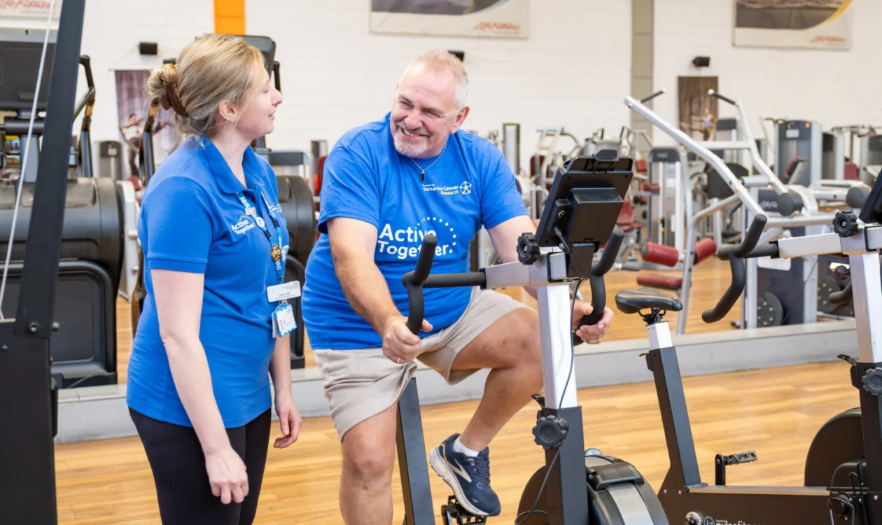 A man on a bike in a gym supported by a woman. Both are in blue t-shirts