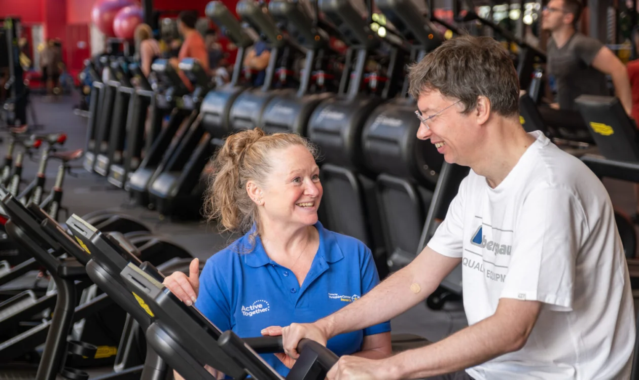 A man on a bike in a gym being supported by a member of staff