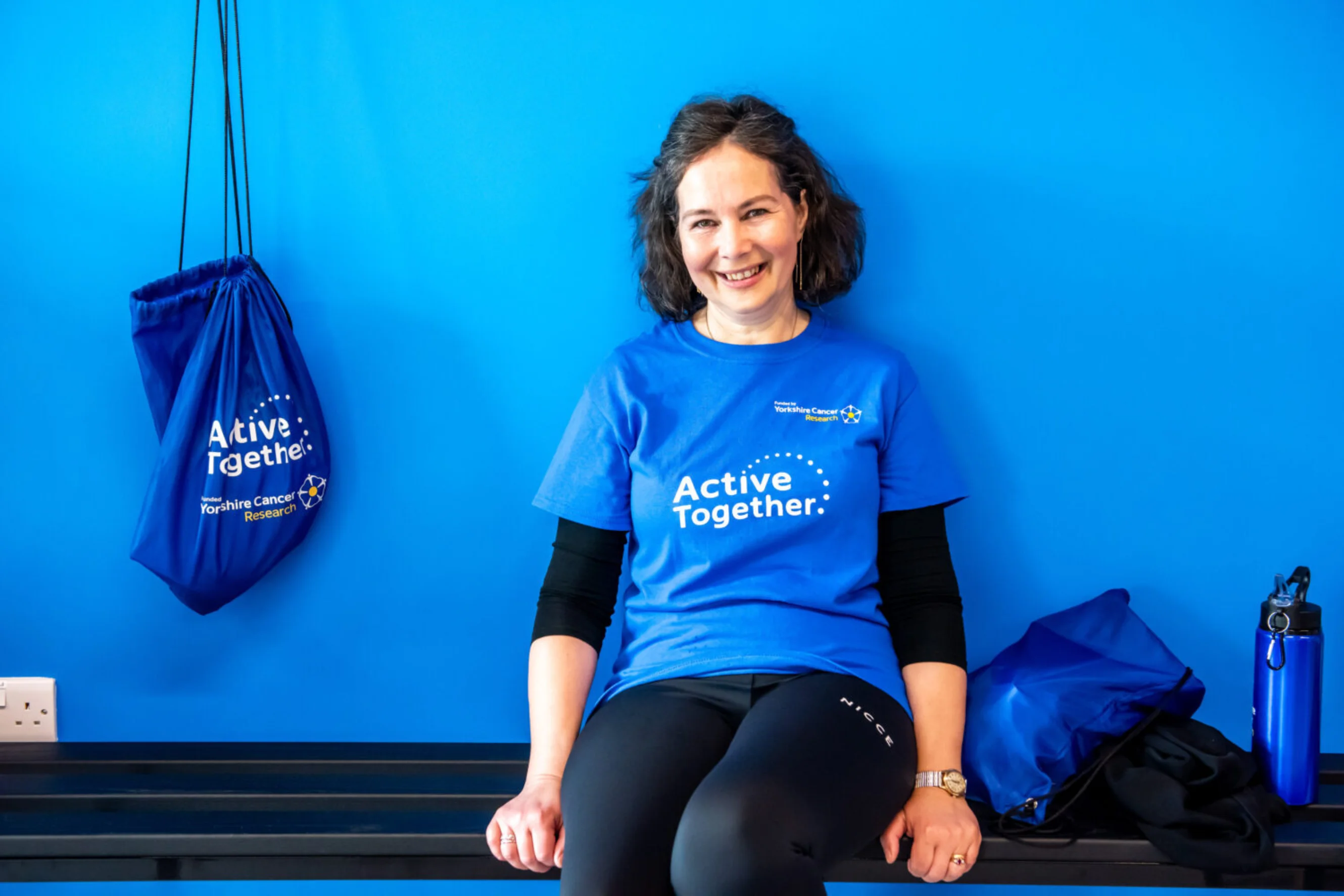 A lady sits on a gym bench with gym kit beside her