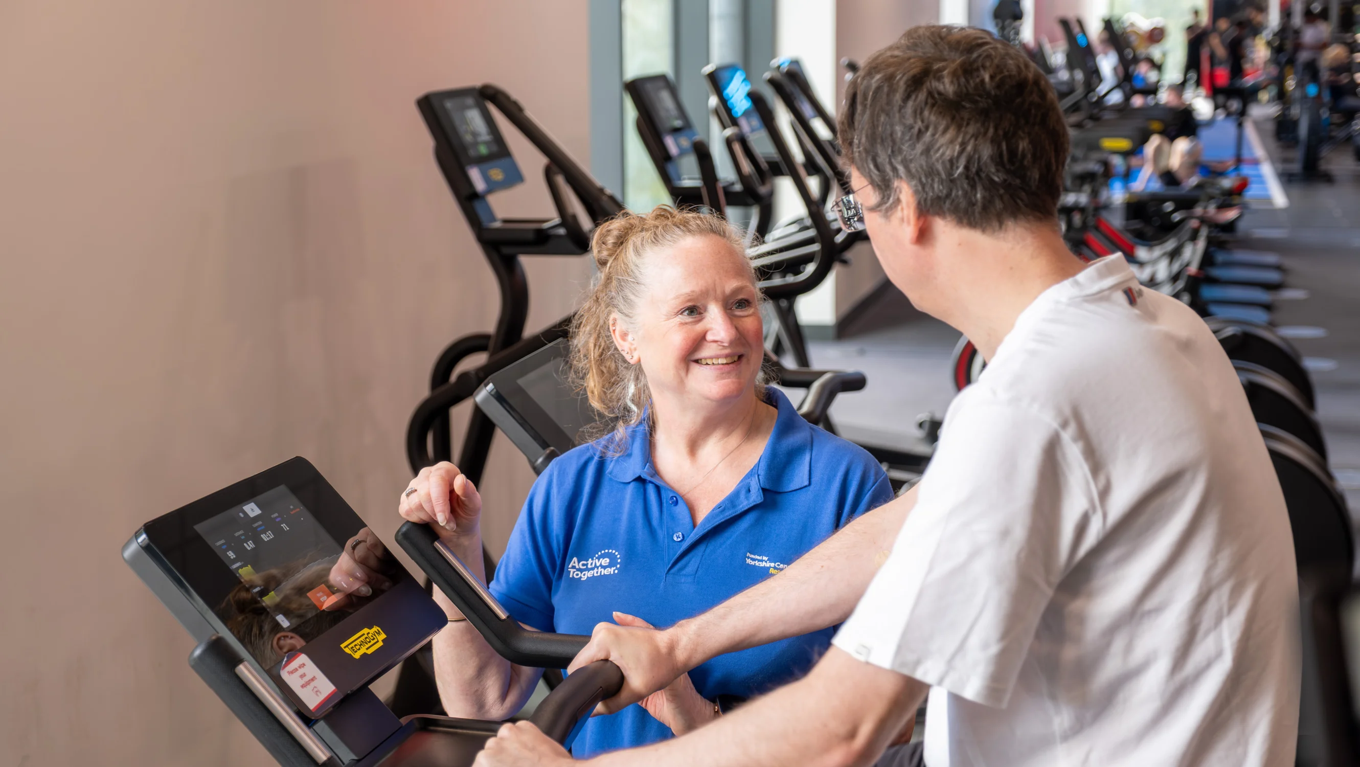 A person exercising on a bike supported by a member of staff in blue