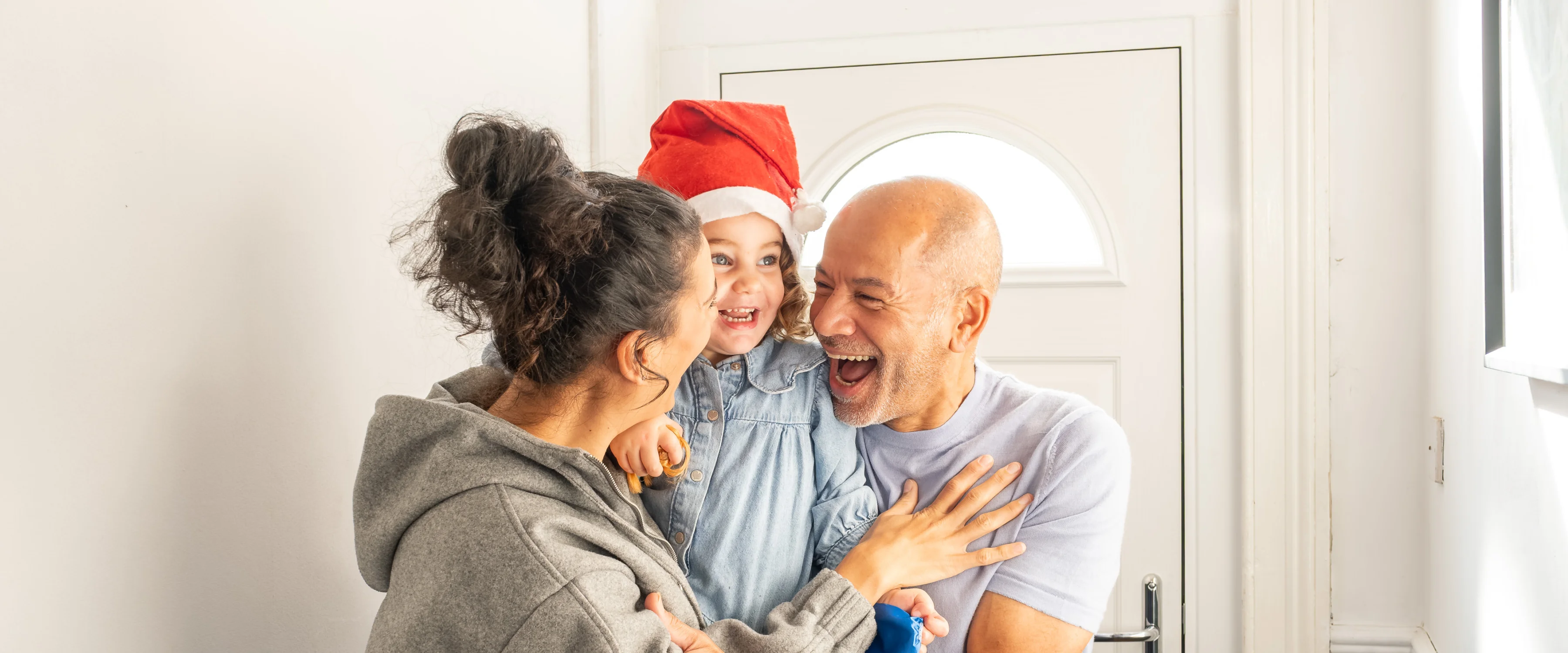 A lady holding a girl in a Santa hat is greeted warmly by a smiling man