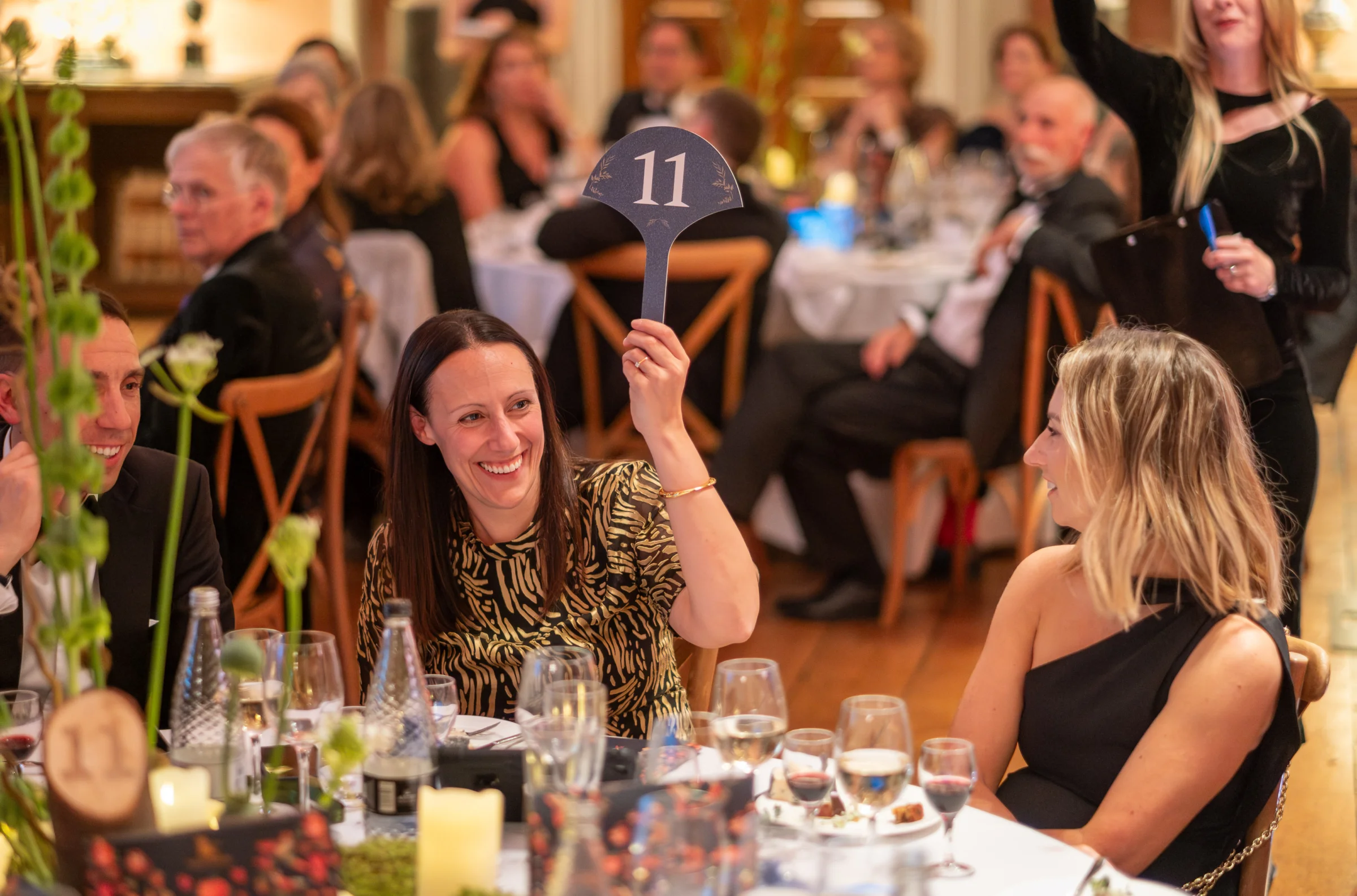 A lady sat at a table at  black tie event raises an auction paddle
